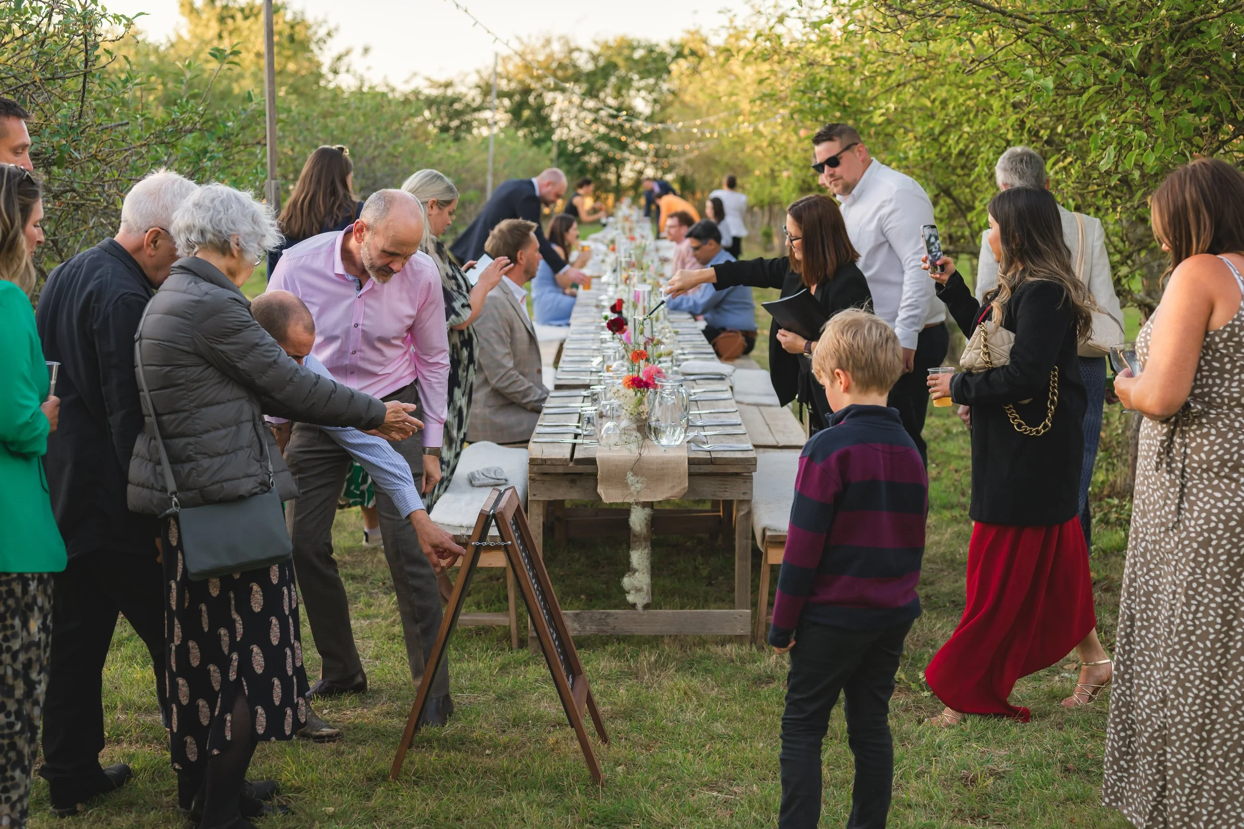 People gathered around a long outdoor table with flowers, enjoying a social event in a green, leafy setting.
