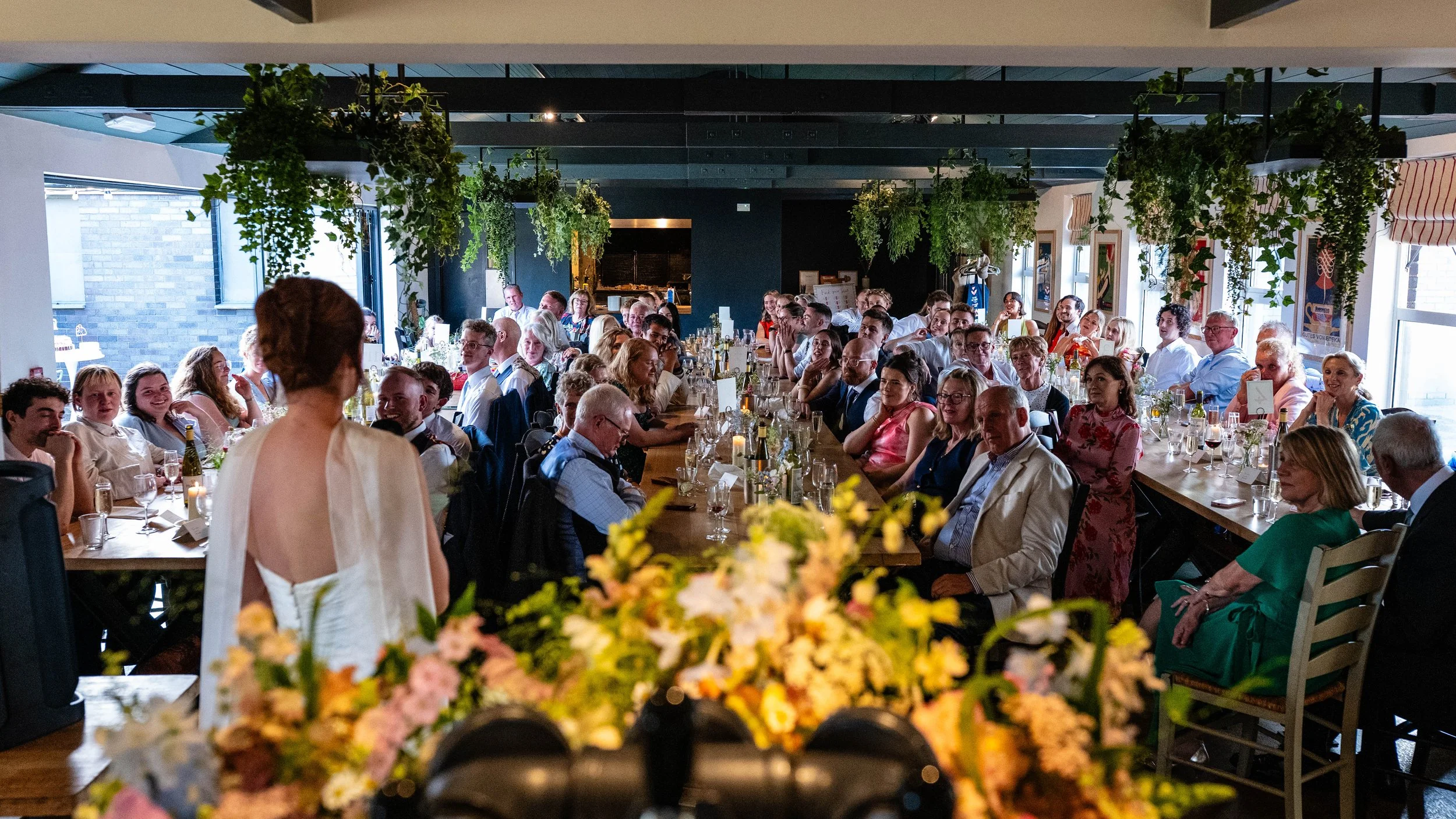 Guests seated at long tables in a restaurant, celebrating an event, with a woman in a white dress speaking, and floral decorations in the foreground.