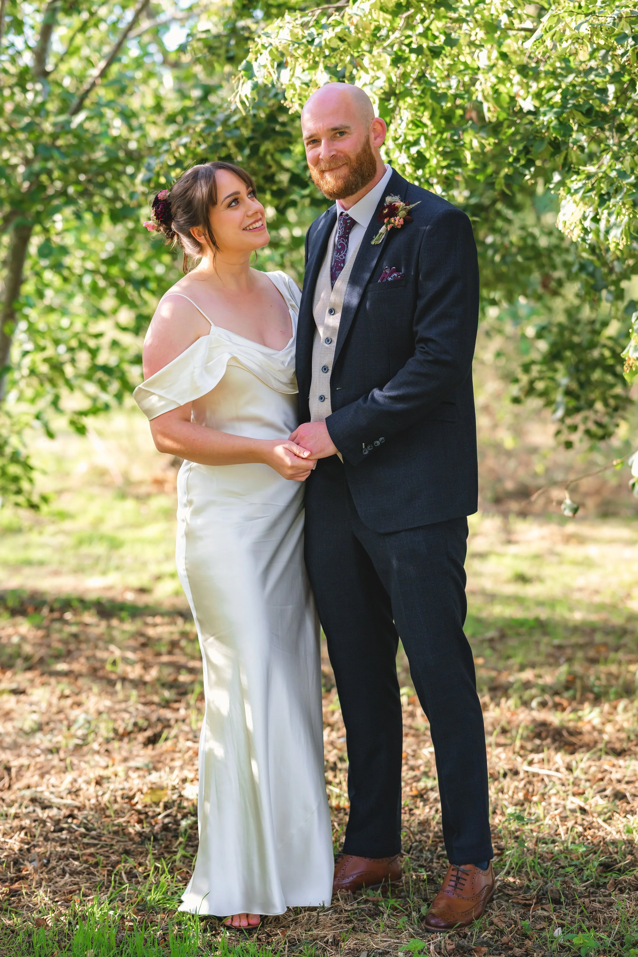 A bride and groom holding hands and standing close together outdoors among green trees and sunlight on the ground.