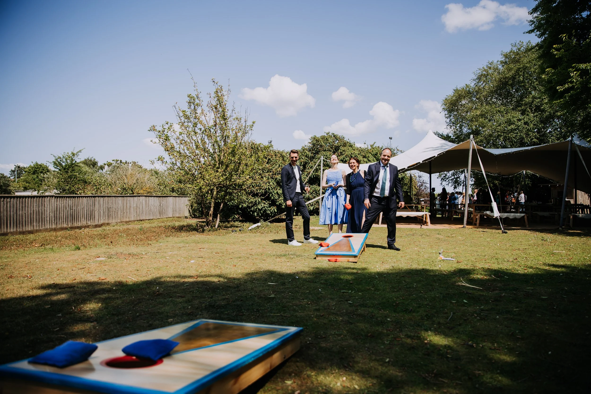 Four people in formal attire playing cornhole outside under a partly cloudy sky, with a tent and trees in the background.