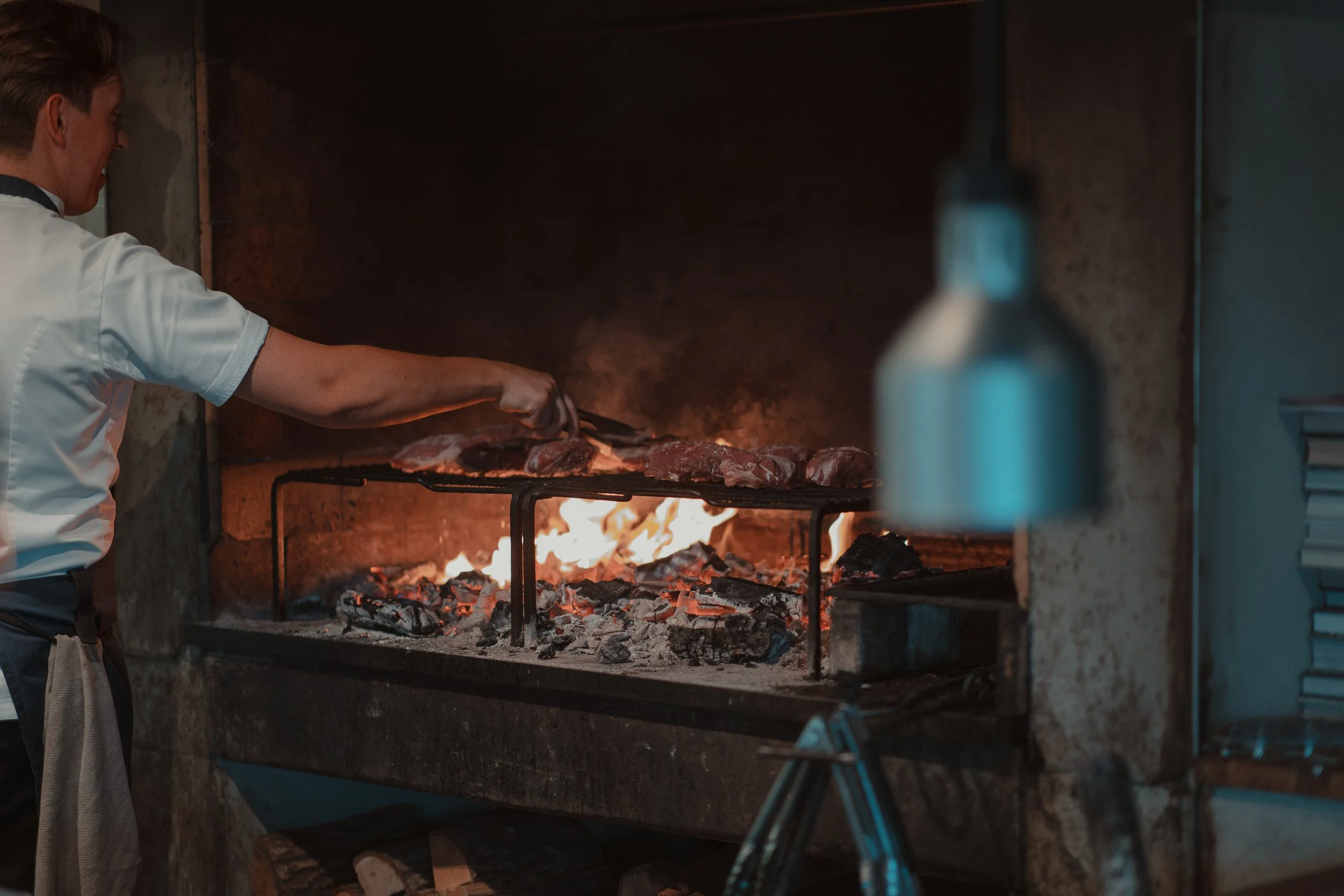 Chef grilling meat over an open flame in a brick oven.