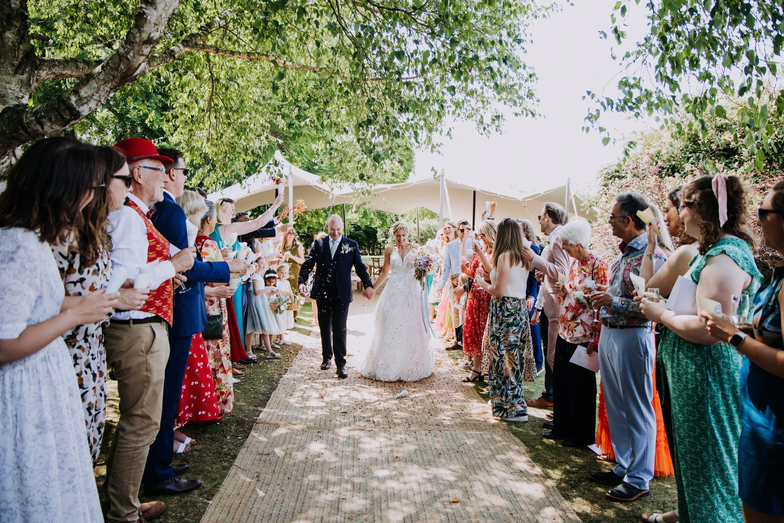 Bride and groom celebrating after their wedding ceremony outdoors under a canopy, surrounded by friends and family holding papers and winceing, with trees and sunlight in the background.