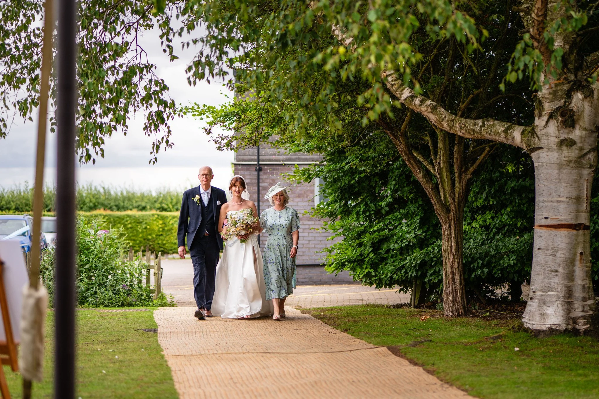 A bride and her parents walking on a path during a wedding, surrounded by trees and greenery, with a cloudy sky overhead.