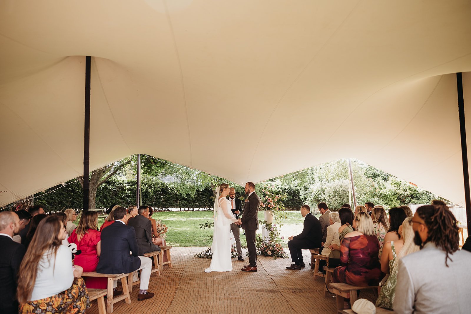 A wedding ceremony taking place under a large tent with couples exchanging vows. Guests seated on wooden benches observe. The scene is outdoors with green trees and a lawn in the background.