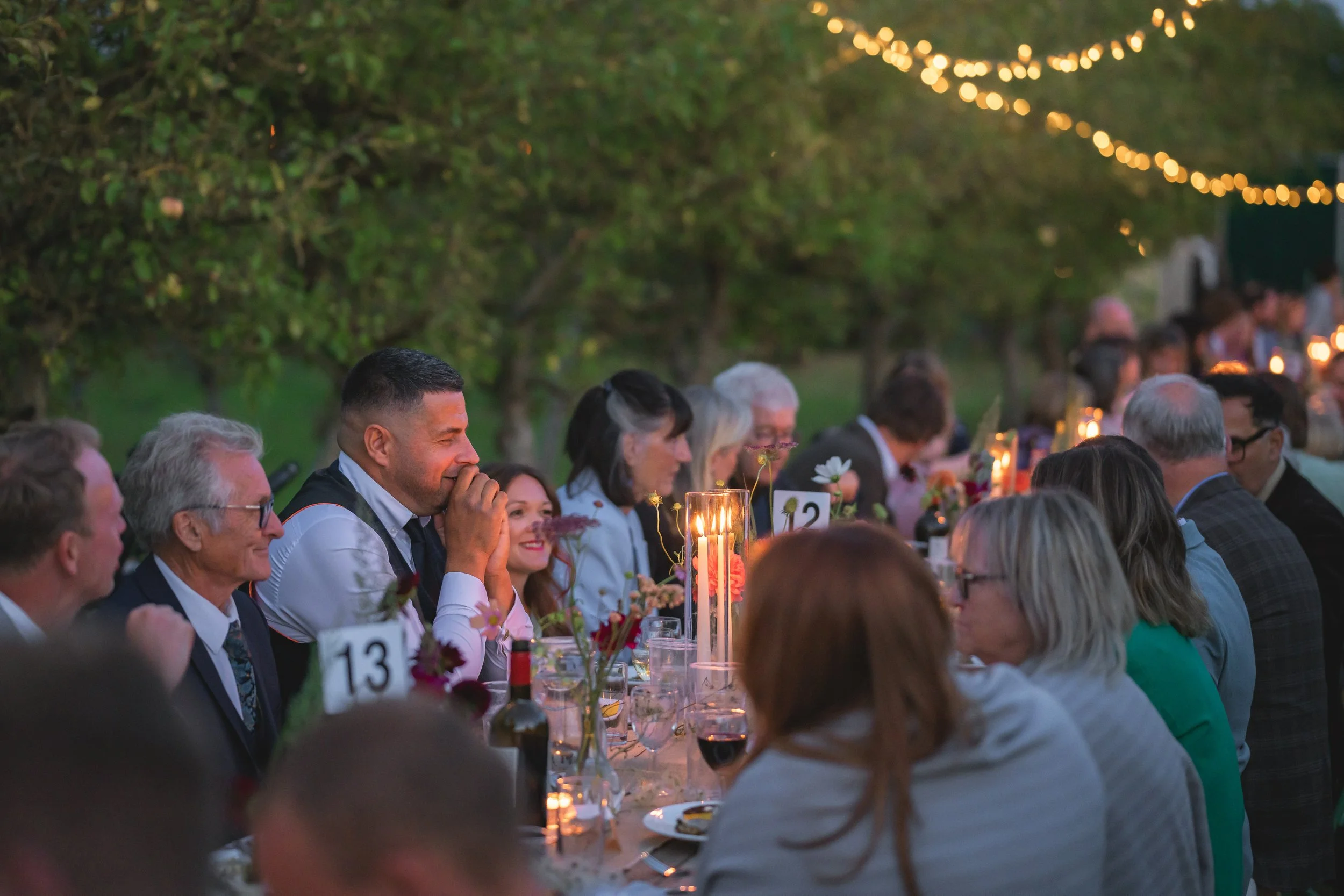 People seated at a long dinner table outdoors during evening, illuminated by string lights, with some guests smiling and others in conversation.