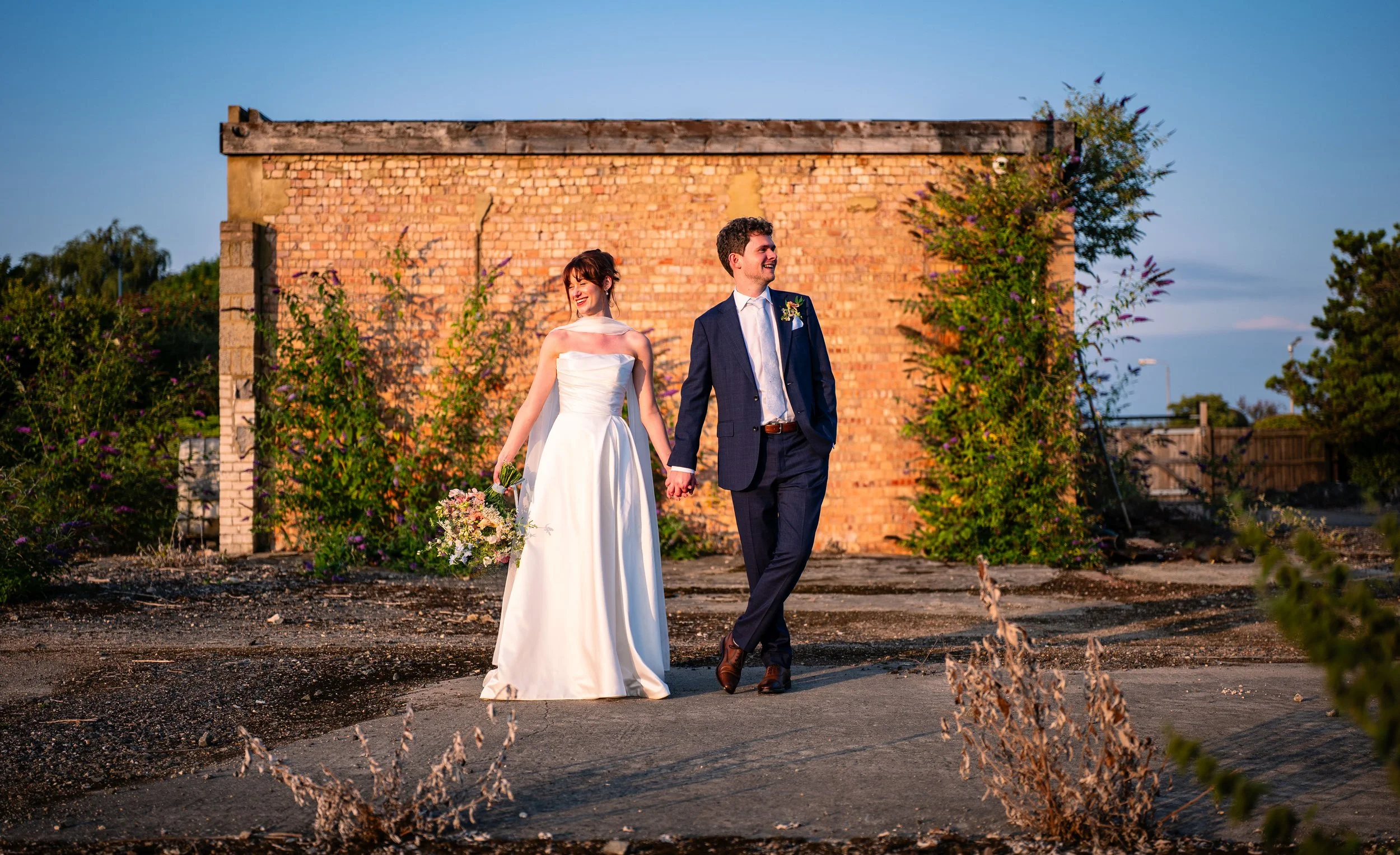 A bride and groom holding hands and walking outdoors at sunset, smiling, with a red brick wall and foliage in the background.