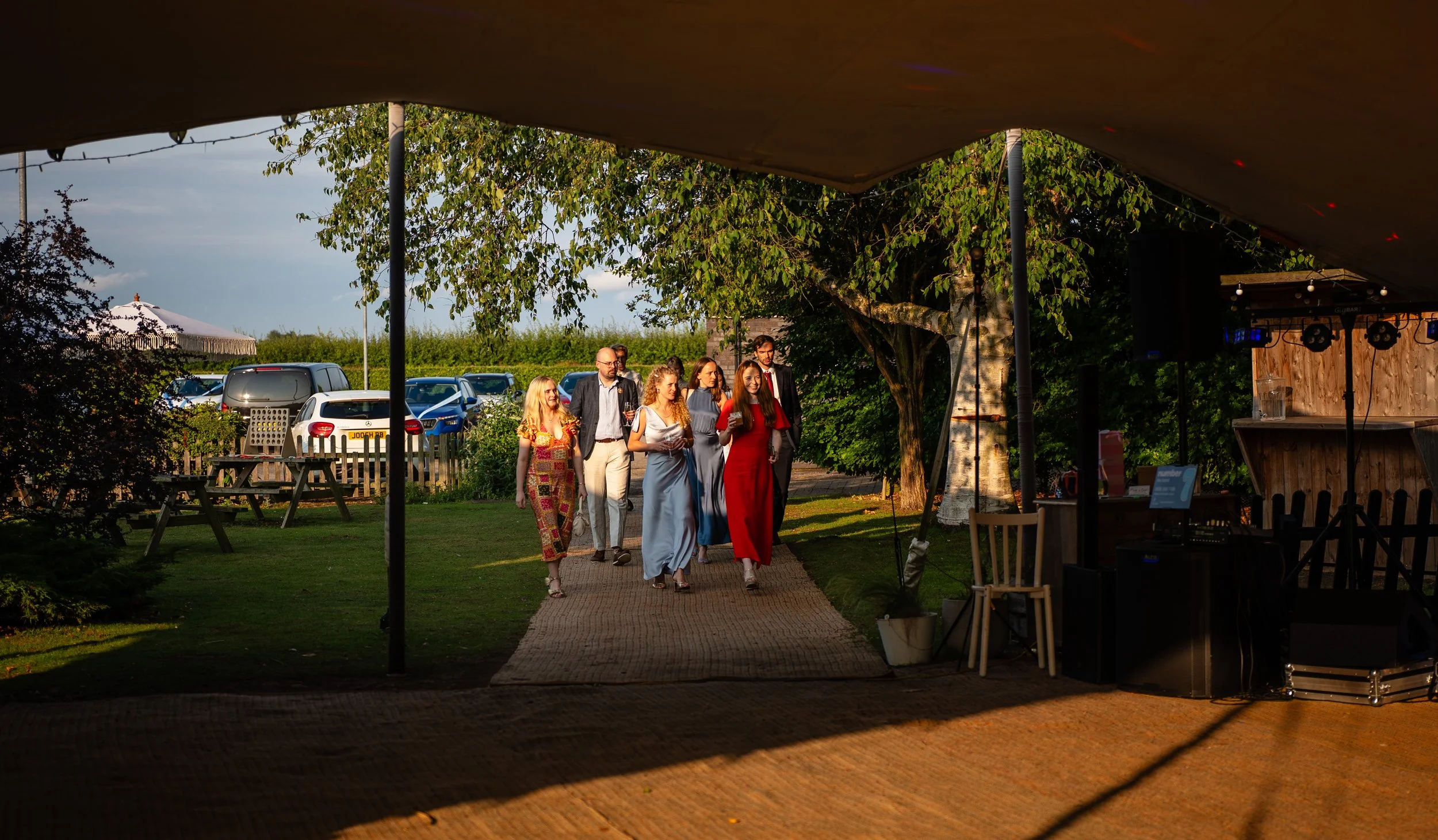 Group of people walking on a pathway outside at sunset, near parked cars and trees, under a tent.