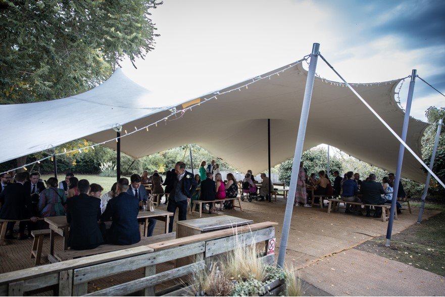 Outdoor gathering of people under a large beige canopy in a park setting with trees and open sky.