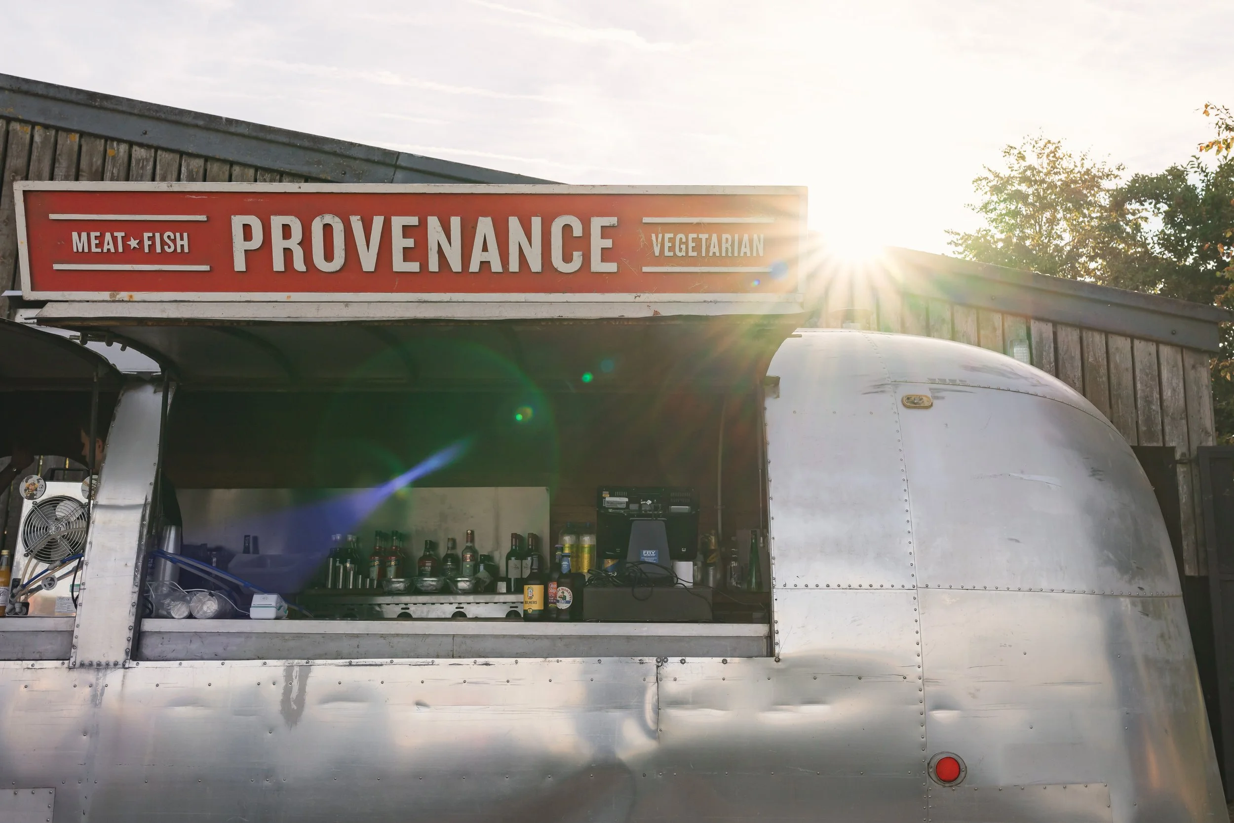 A food truck with a sign reading 'Provenance Vegetarian' and 'Meat Fish,' with bottles and a cash register visible inside, parked in front of a wooden building with trees and the sun shining in the background.