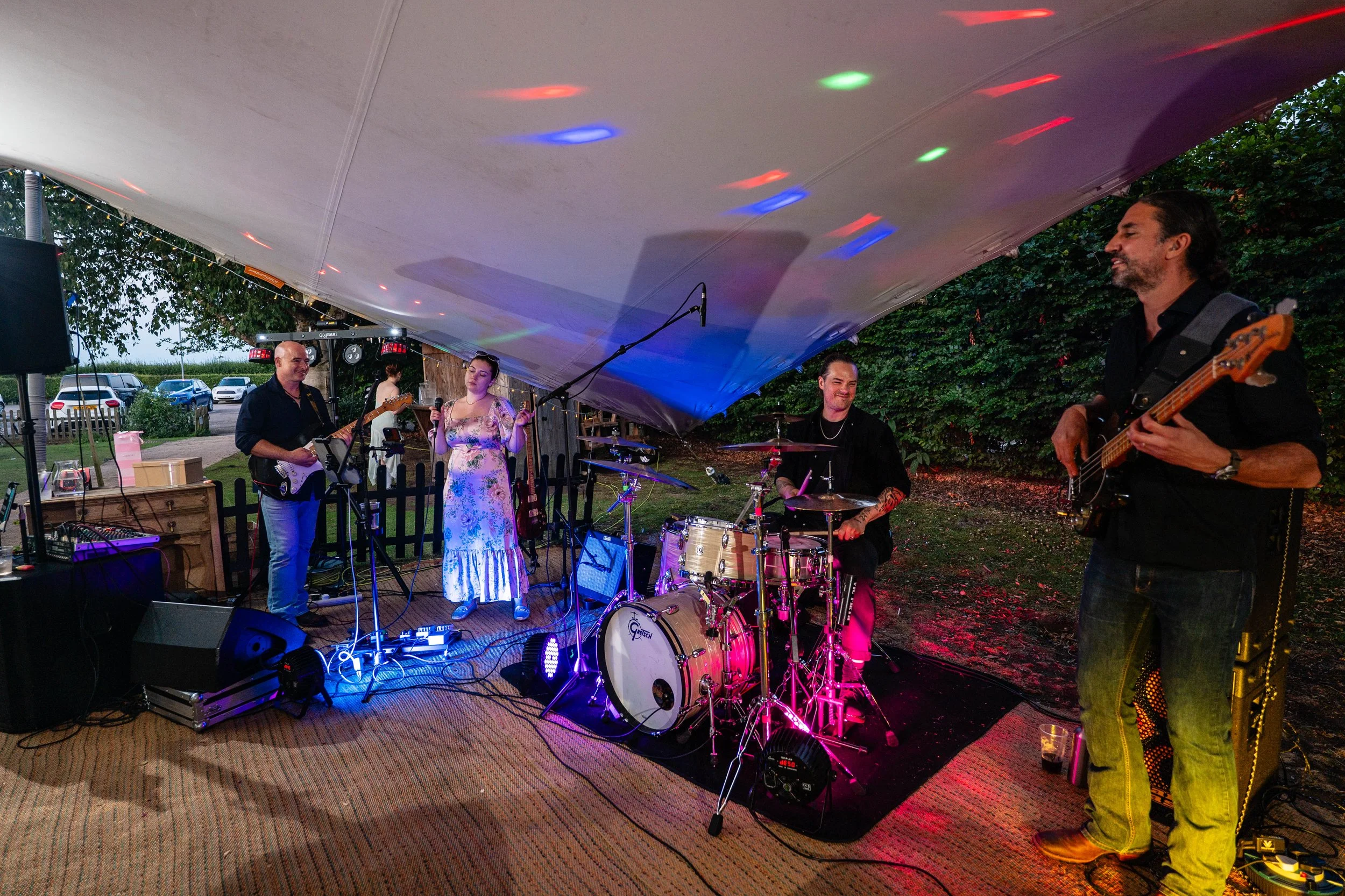 A band performing outdoors under a tent with colored lights, including a guitarist, a female vocalist, a drummer, and a bassist, with parked cars and trees in the background.