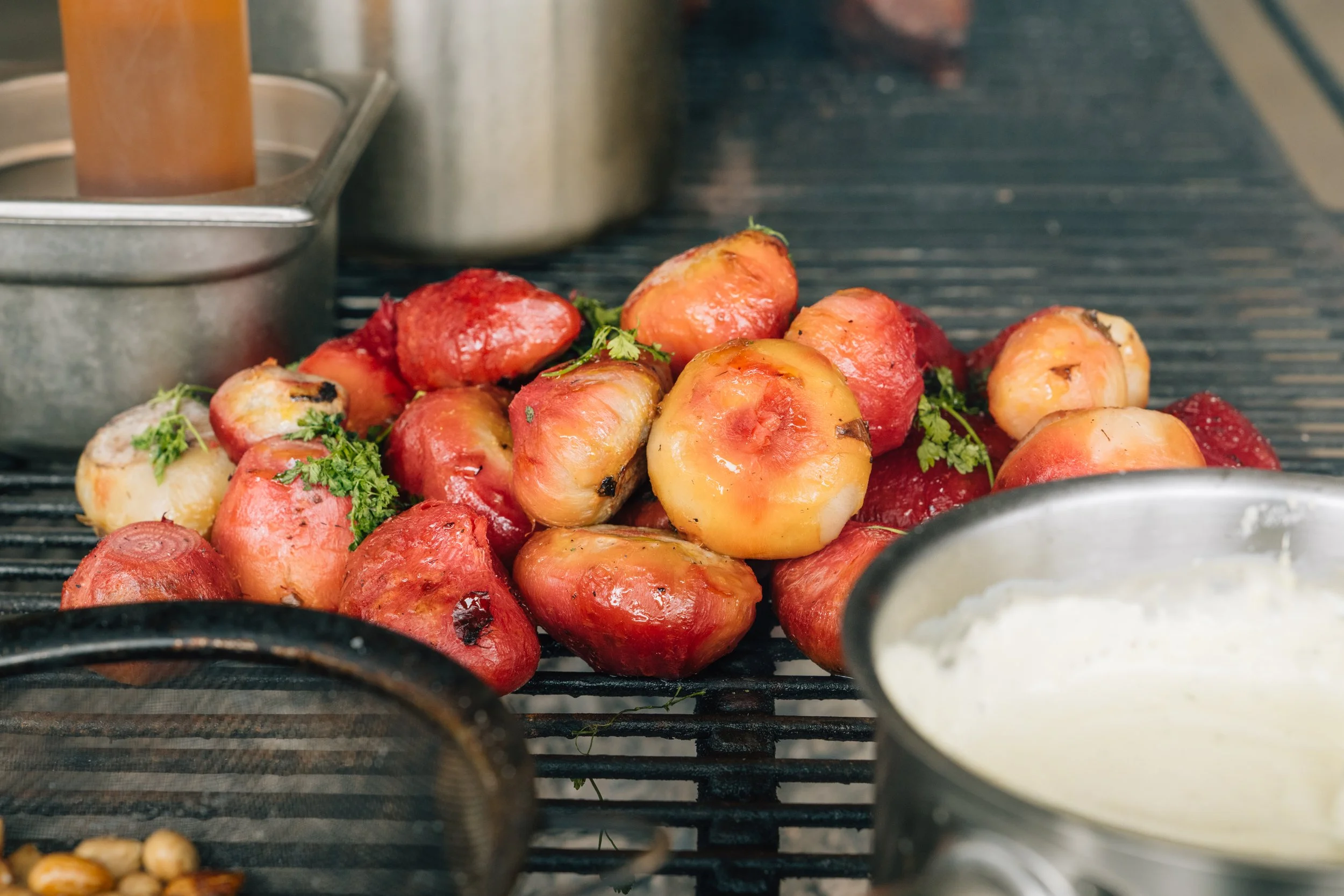 Charred tomatoes and herbs on a grill, with a pot of creamy sauce in the foreground.