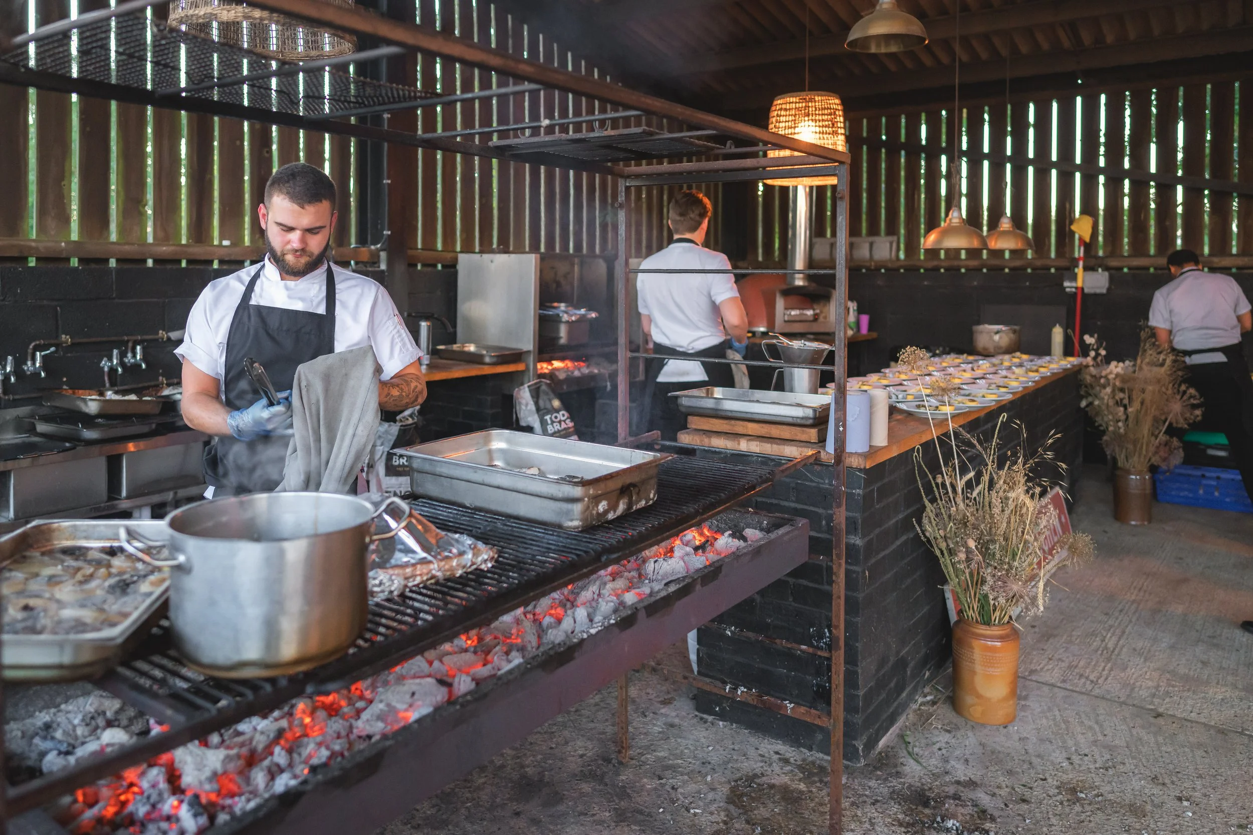 Chefs preparing food in an outdoor kitchen with a rustic, open-air setting, including a grill over hot coals, cooking utensils, and numerous prepared dishes on the counter.
