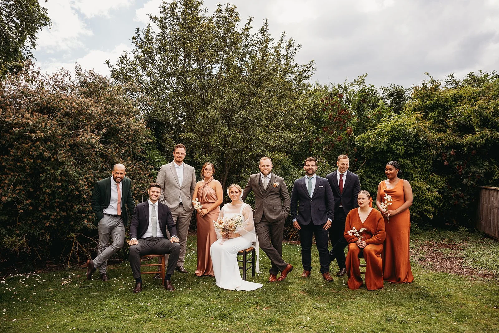 Group of people at a wedding outdoors, with trees and cloudy sky in the background. The bride is seated in the center holding a bouquet, and the groom is standing next to her. Bridesmaids and groomsmen are standing and sitting around them, dressed in