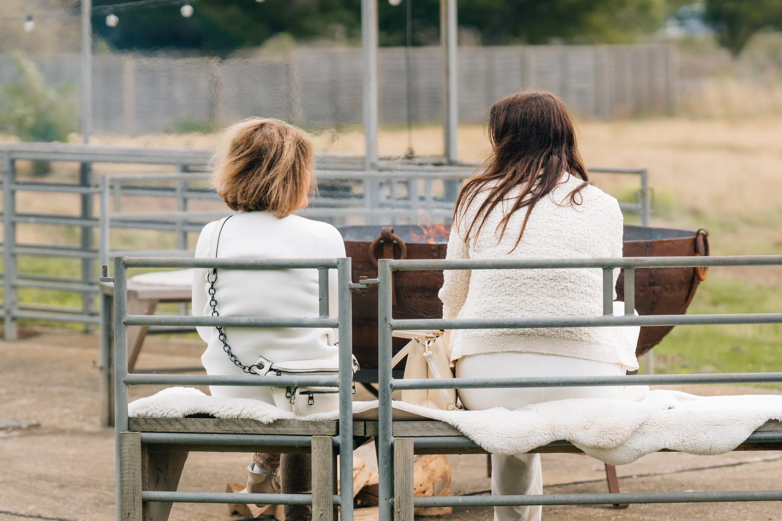 Two women sitting on a bench near a fire pit, looking away from the camera, in an outdoor setting.