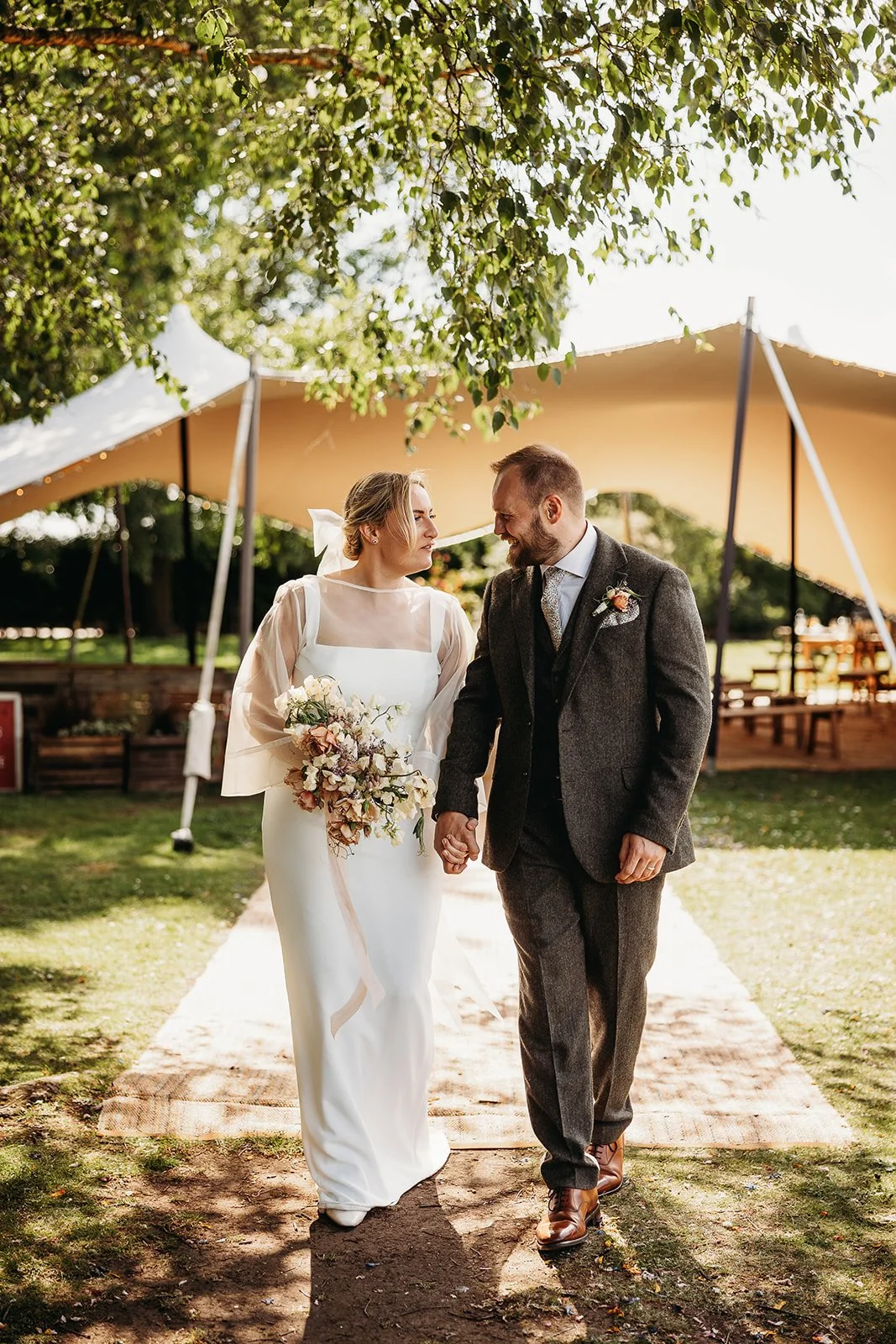 A bride and groom walking hand in hand outdoors under a large trees and a beige tent, smiling at each other on their wedding day.
