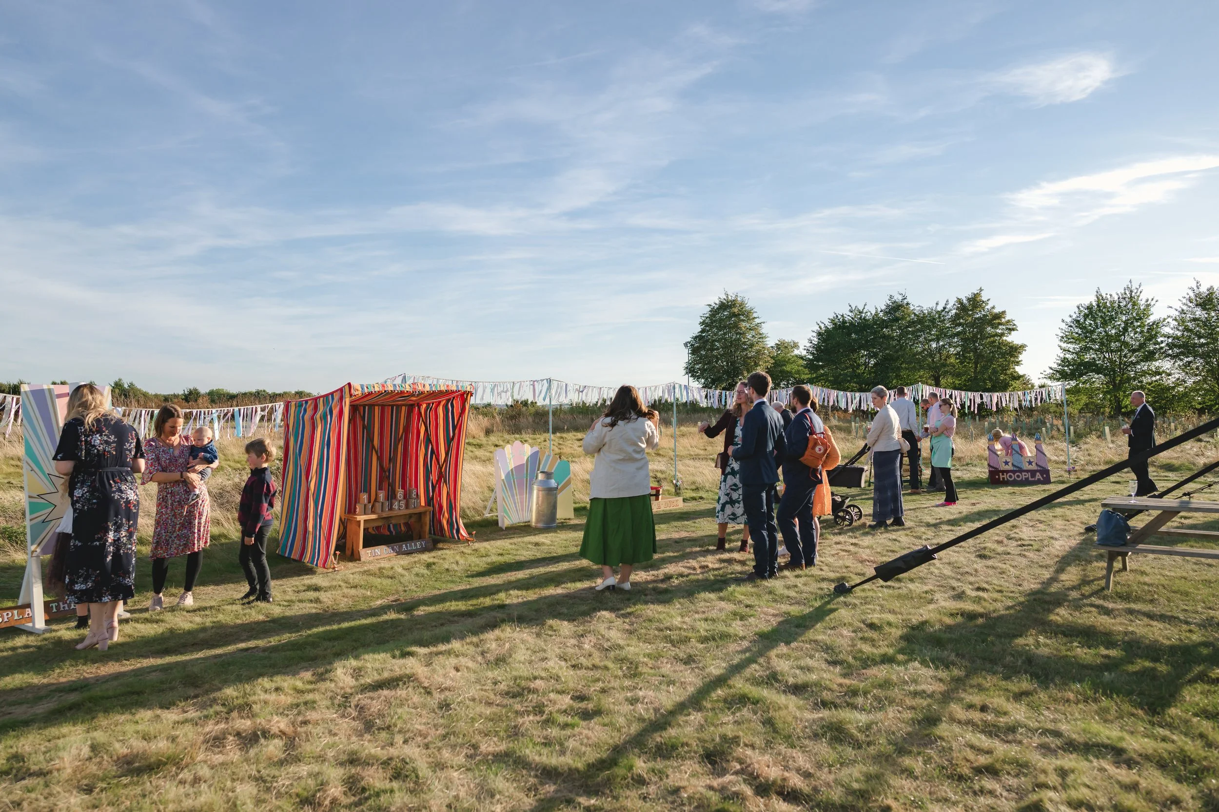 Group of people at an outdoor fair or market on a sunny day, with colorful stalls and decorations, grassy field, blue sky, trees in the background.