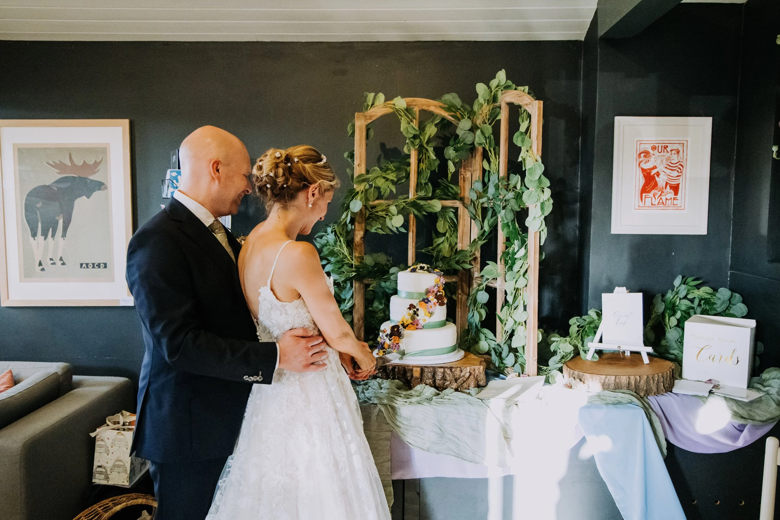A bride and groom cutting a wedding cake decorated with flowers, standing beside a table with wedding cards and decorations, in a room with dark walls and framed artwork.