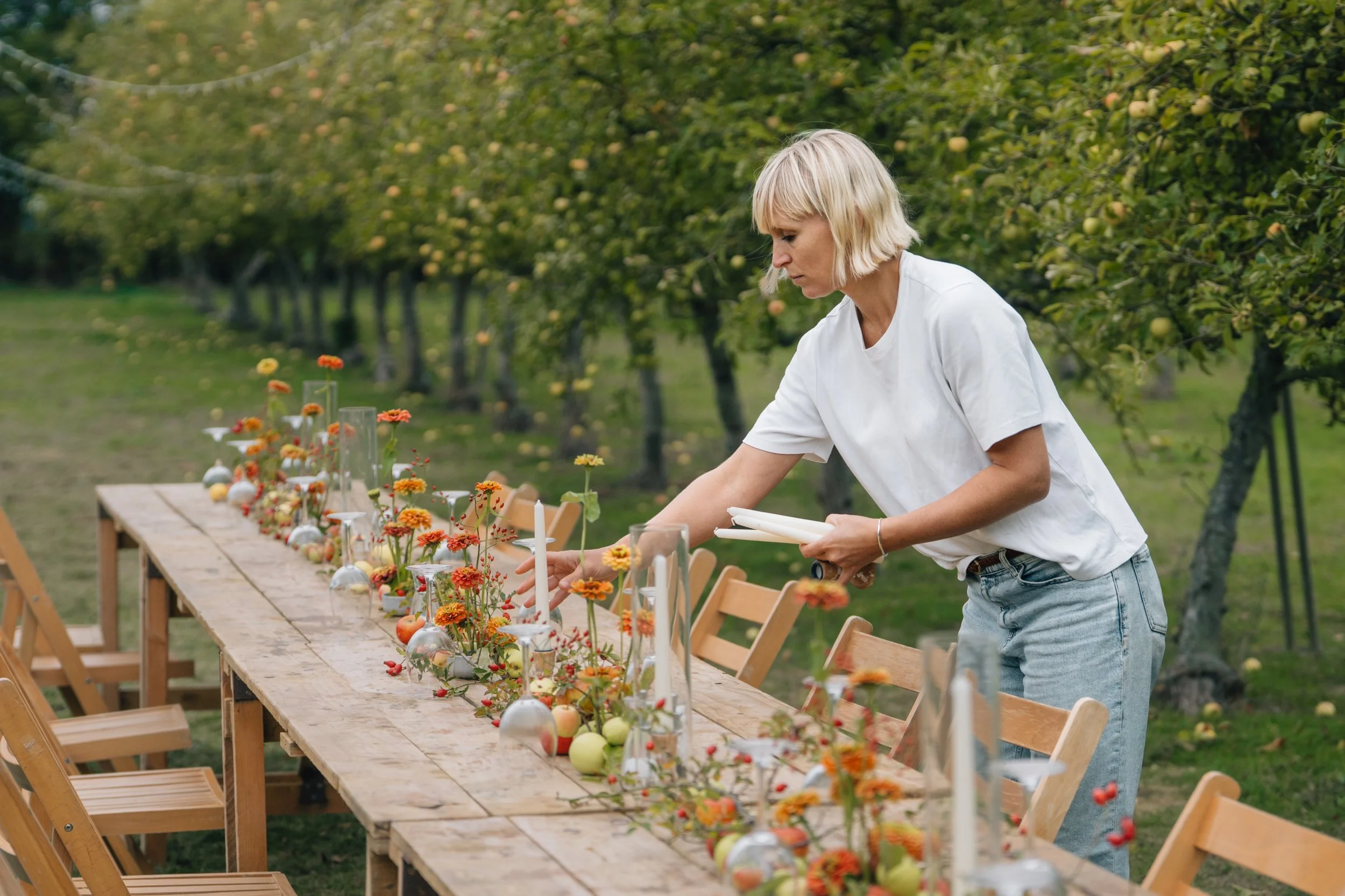 A woman is decorating a long wooden table outdoors with flowers, candles, and apples, amidst a backdrop of green trees and a clear sky.