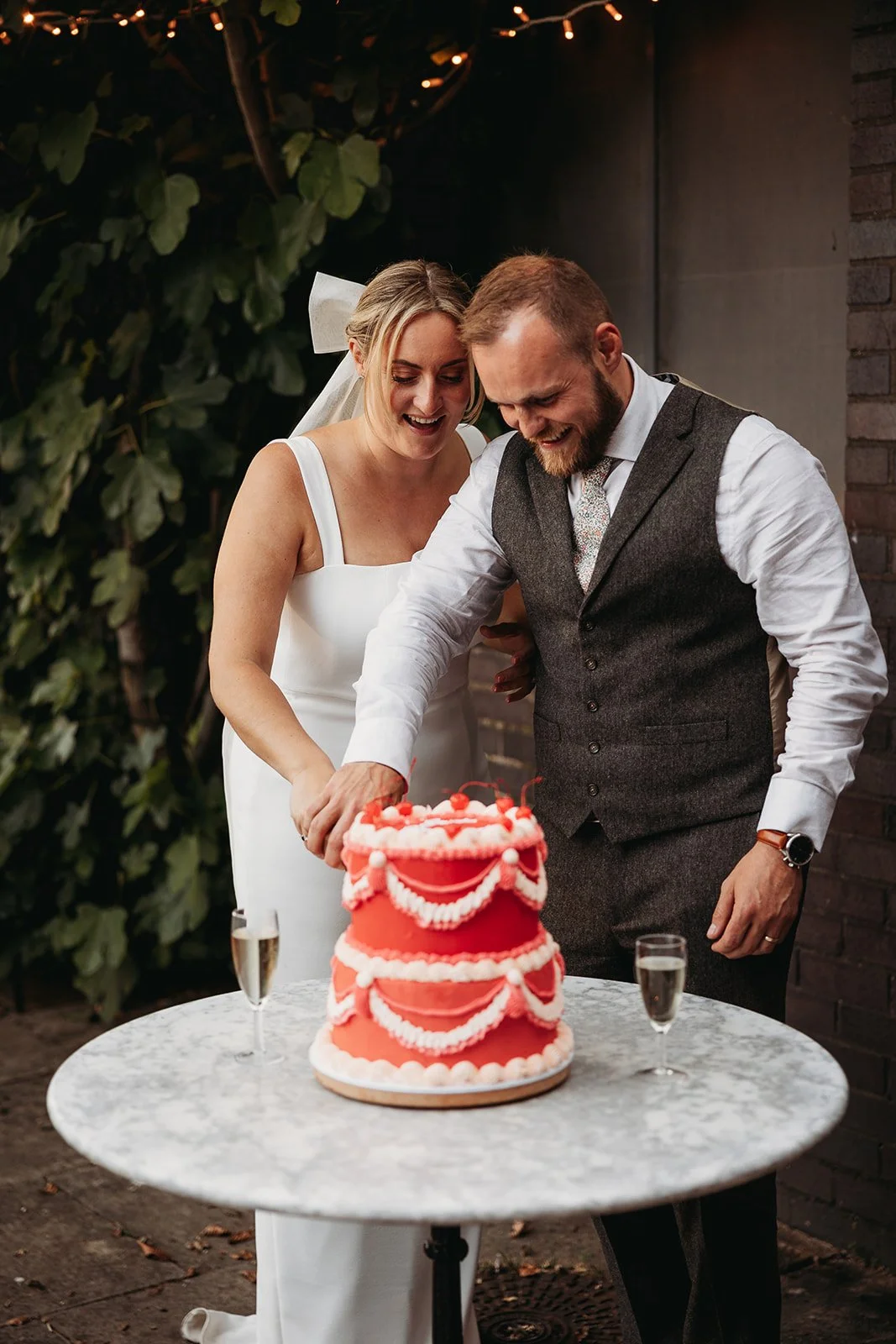Bride and groom cutting a wedding cake at their reception, with champagne glasses on the table and fairy lights overhead.