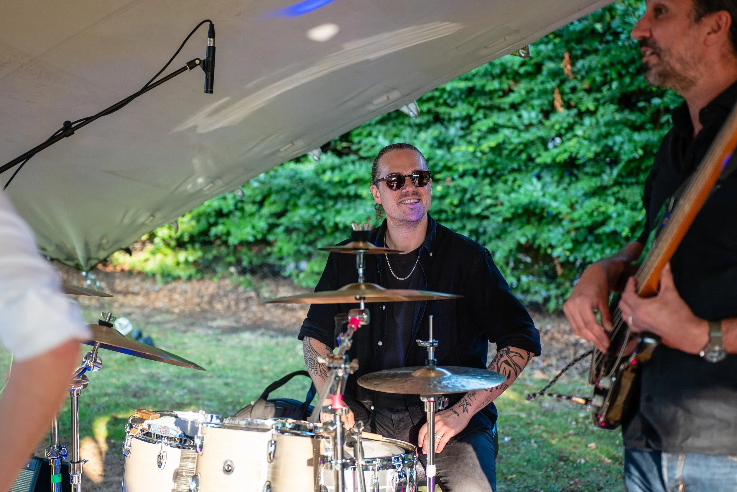A musician wearing sunglasses and black clothing playing drums, with another person playing guitar nearby, outside with green bushes in the background.