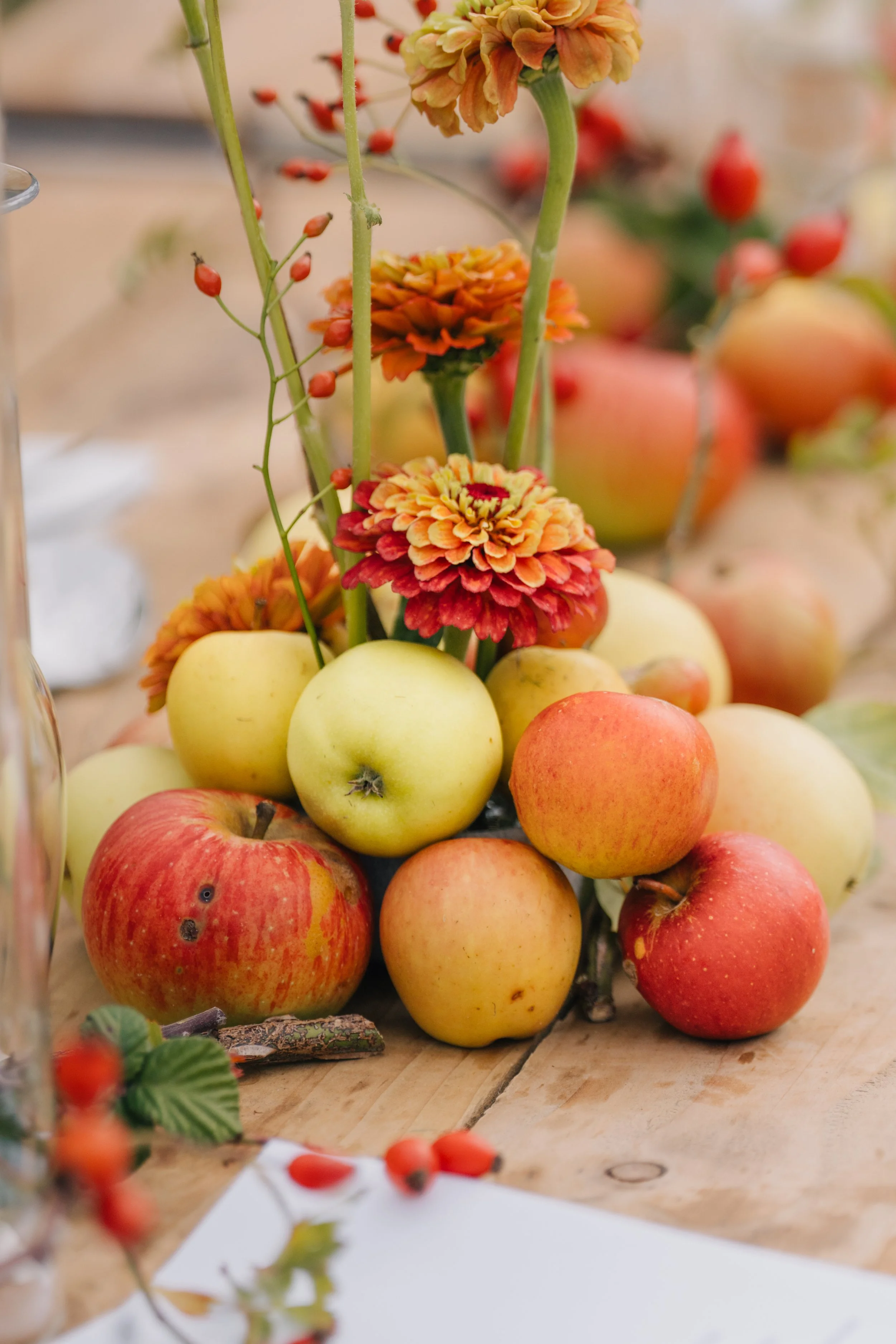 A wooden table with a floral and fruit centerpiece including orange and yellow flowers and red and yellow apples.