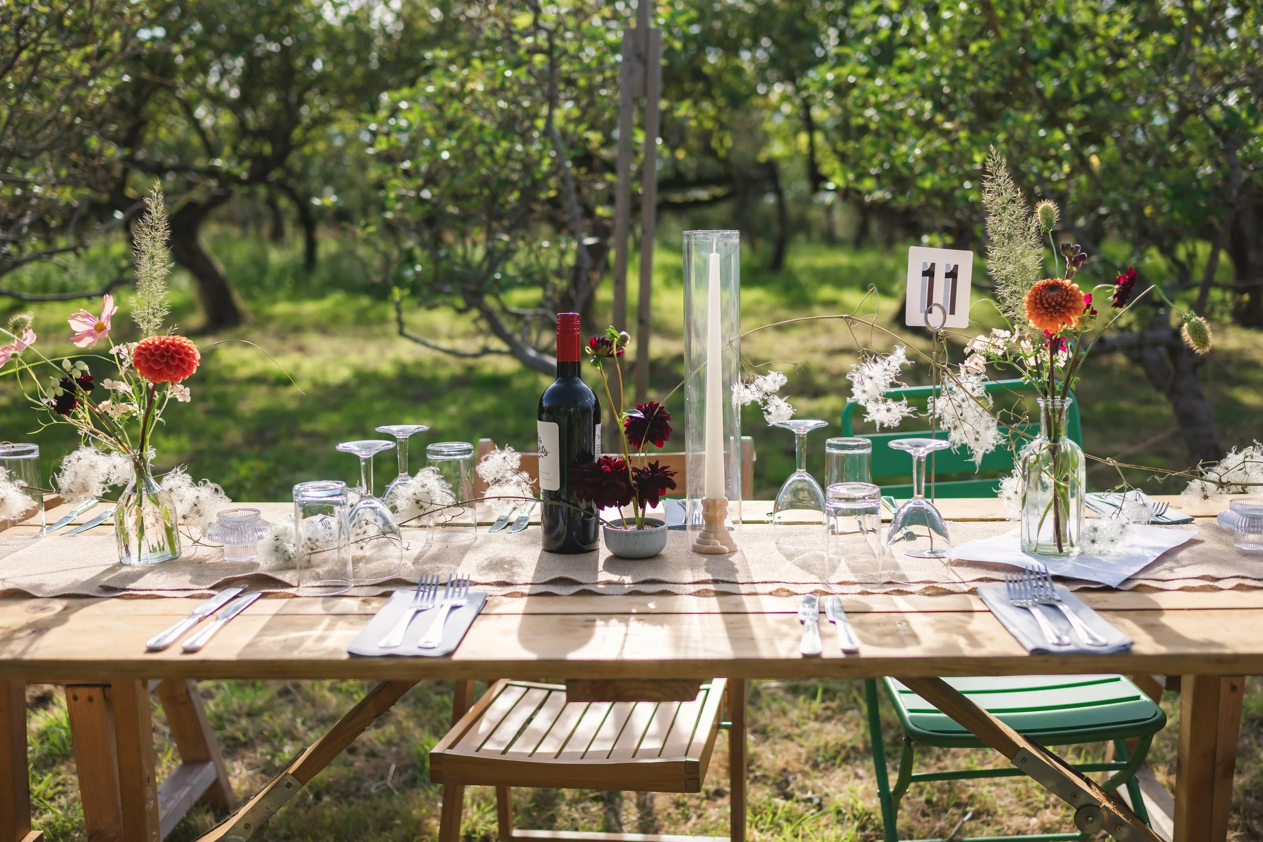An outdoor dining table set in a vineyard with flowers in glass bottles, a wine bottle, a tall candle, and tableware. The table is decorated for a special occasion in a natural, rustic setting.