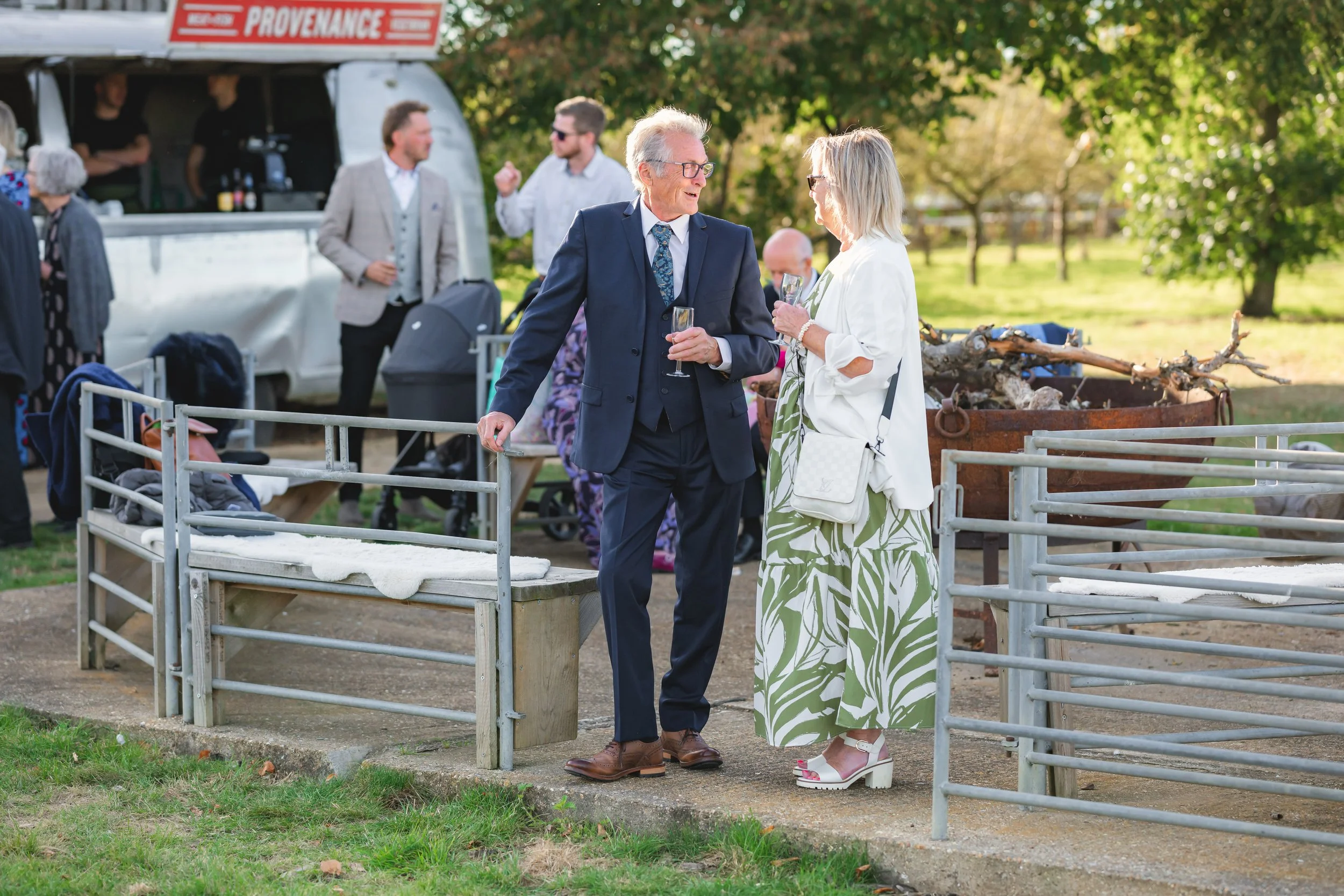 Two elderly people in formal attire talking and holding glasses at an outdoor event, with a food truck labeled "Provenance" in the background and other guests mingling.