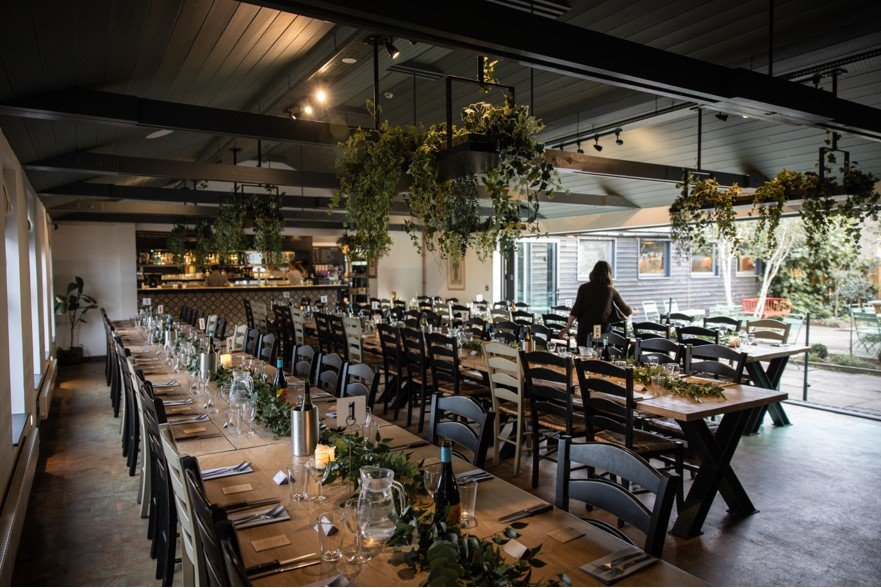 Interior of a restaurant or event space set up for a meal with long tables, wine bottles, glasses, and green foliage decorations, large windows letting in natural light, and a person walking near the window.