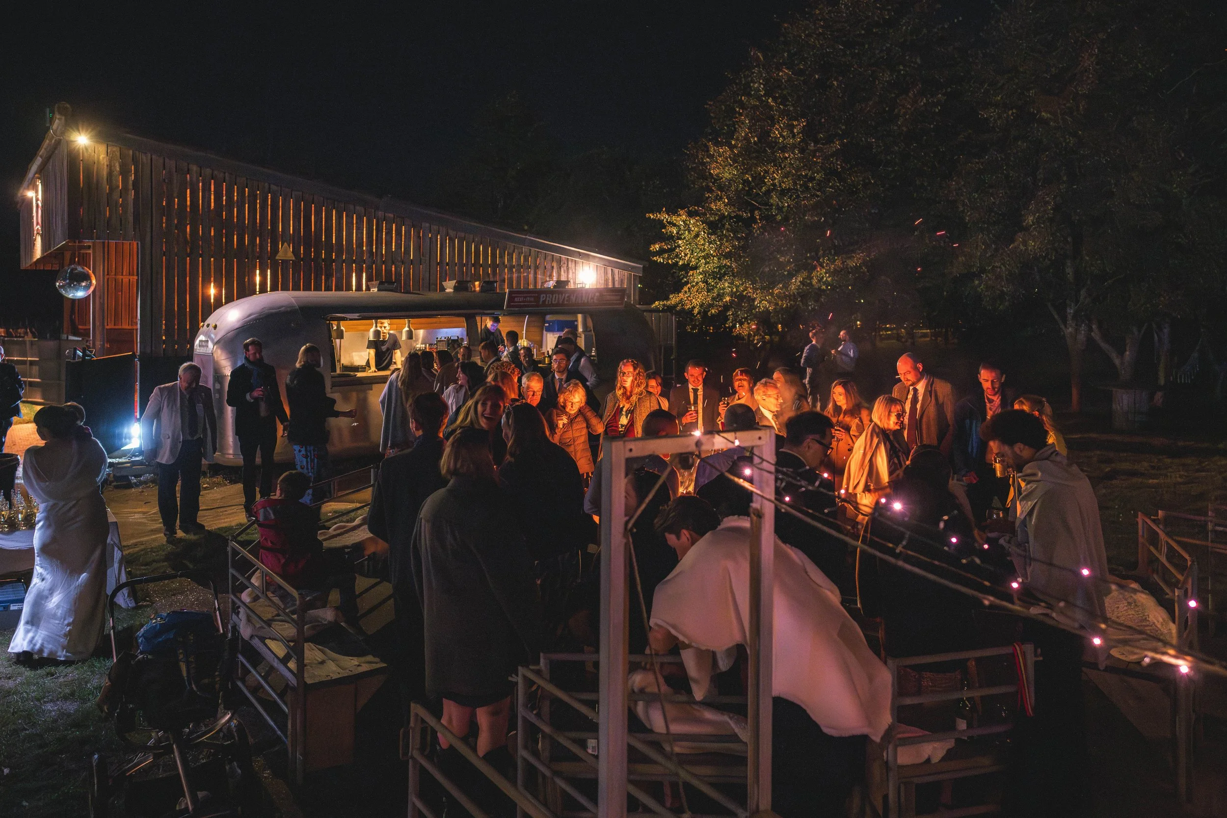 Nighttime outdoor social event with people gathered around a food truck and tables, illuminated by string lights, with trees and a wooden building in the background.