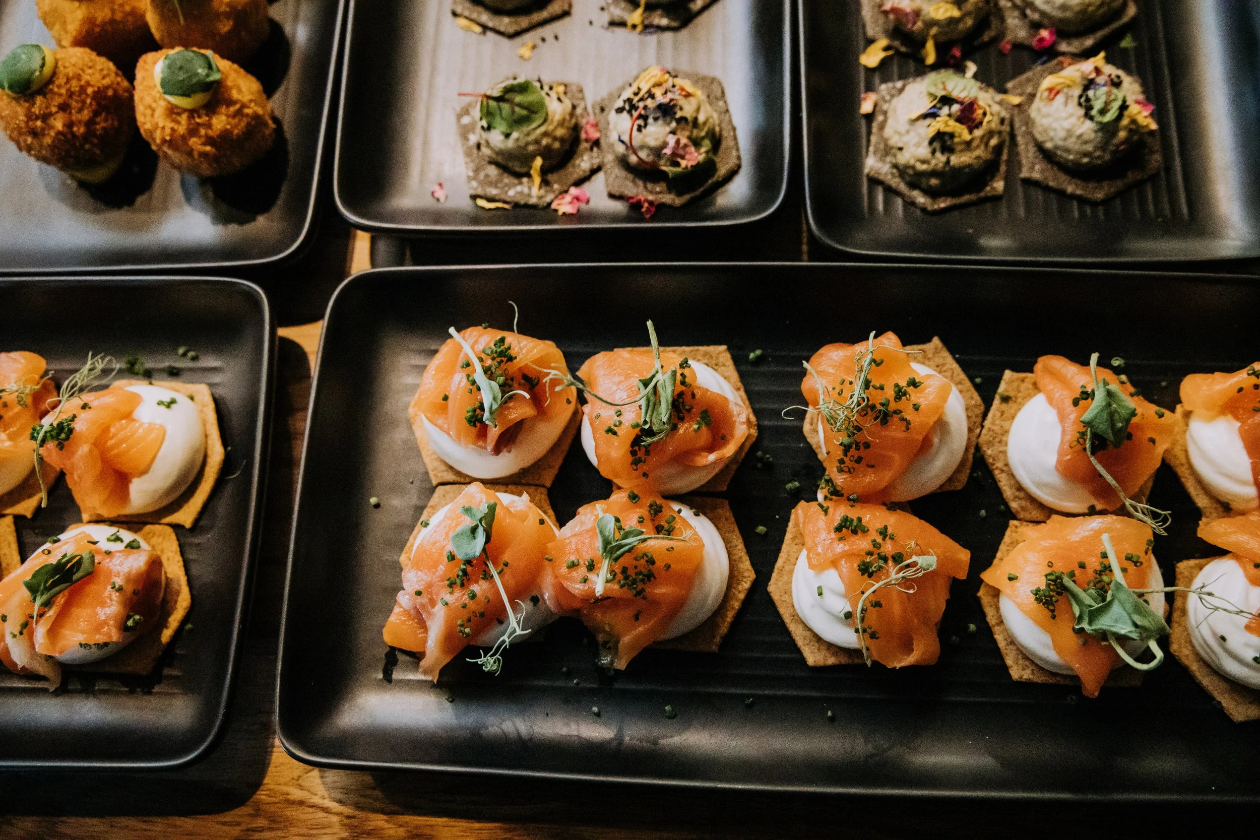 Assorted appetizers on black trays, featuring smoked salmon on crackers with herbs, and various other bite-sized treats garnished with herbs and edible flowers.