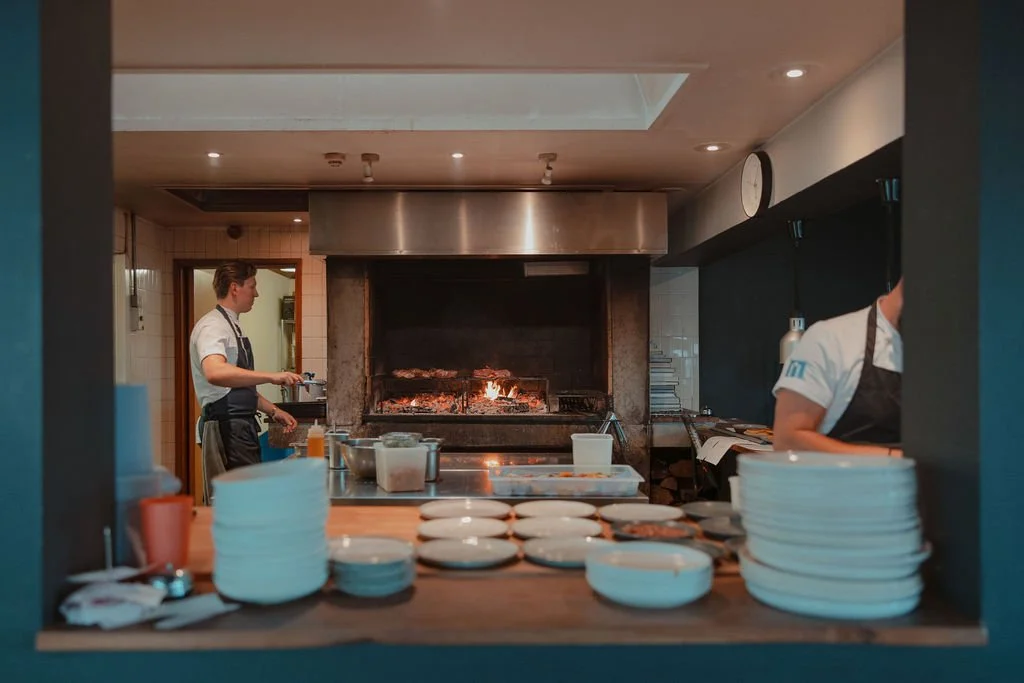 View of a restaurant kitchen with two chefs, a wood-fired grill with flames, stacks of plates, and various cooking utensils.