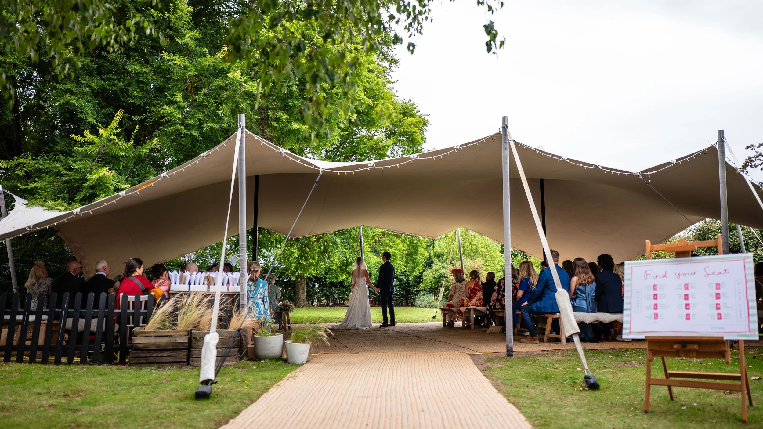 An outdoor wedding ceremony taking place under a large cream-colored canopy tent, with guests seated on wooden benches on either side of an aisle. The bride and groom are standing at the altar holding hands, with the bride in a white dress and long v