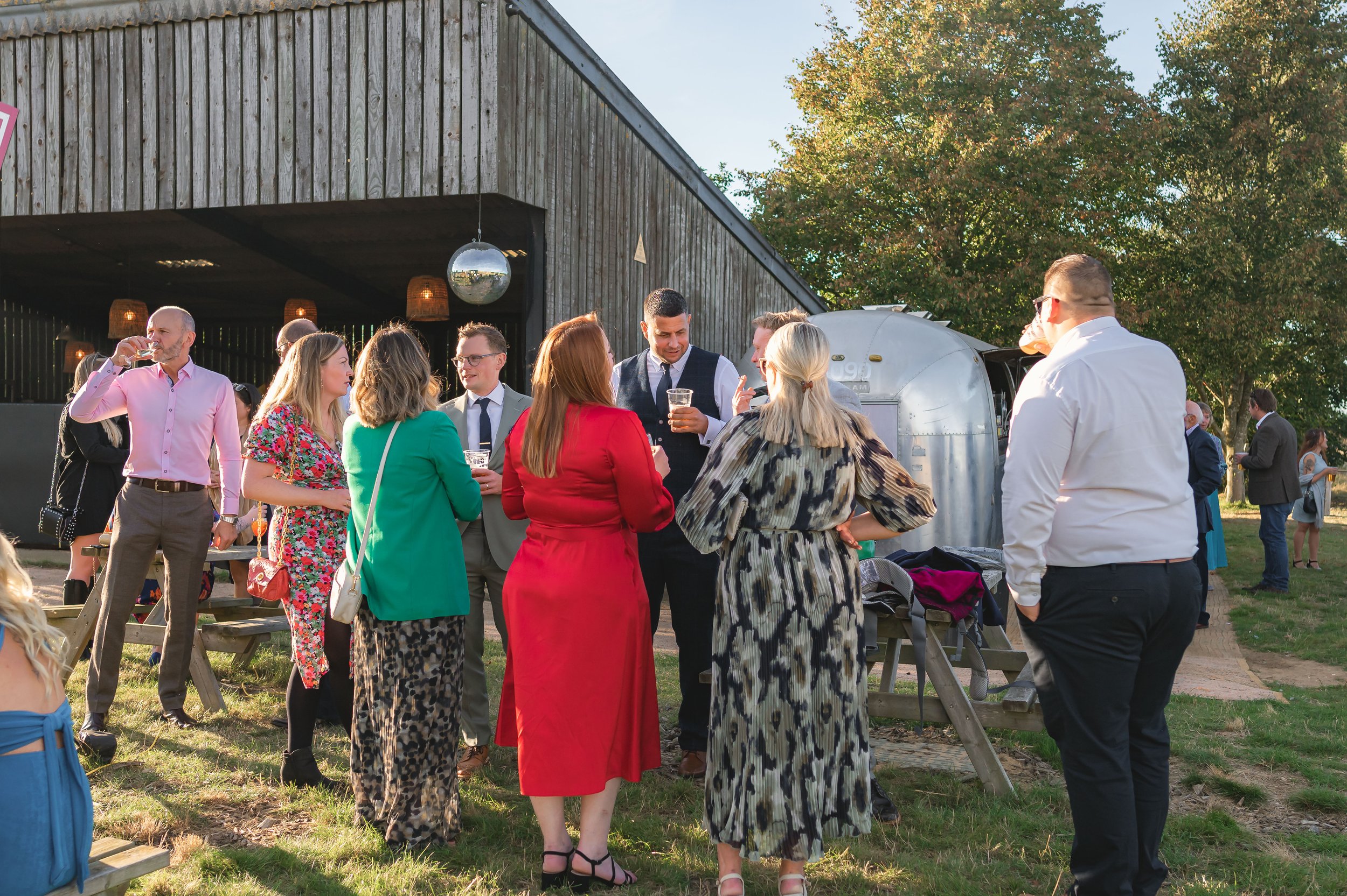 Group of people socializing outdoors near a rustic barn during the evening, some holding drinks, with a wagon and trees in the background.