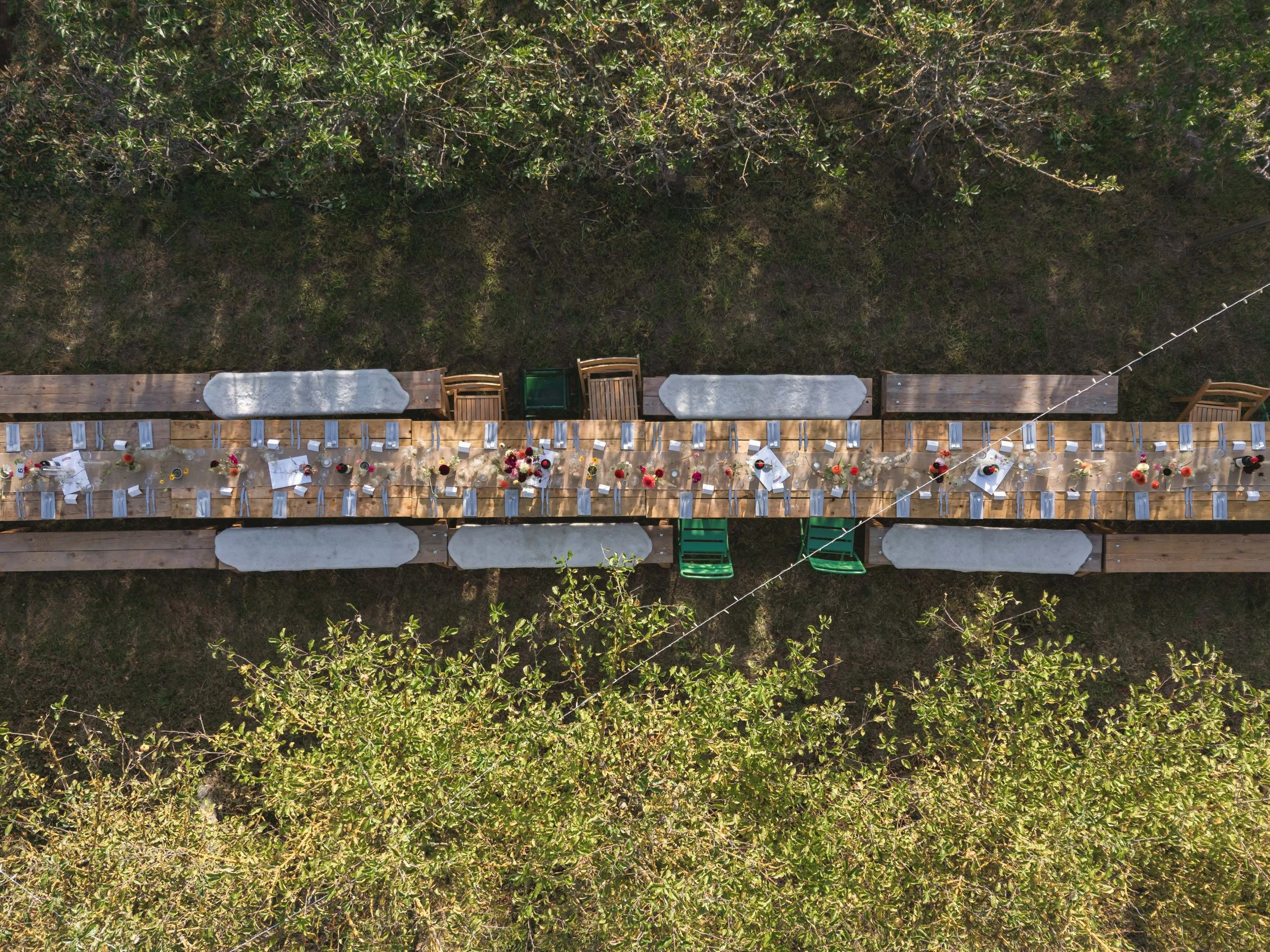 Overhead view of a long wooden outdoor dining table set for a celebration, with chairs, white tablecloths, and floral centerpieces, in a natural outdoor setting with trees and grass.