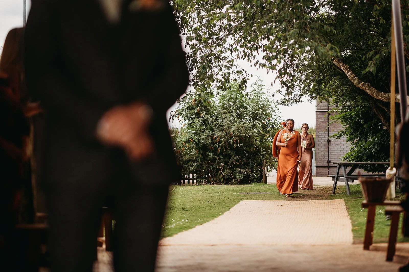 Two women walking on a woven pathway outdoors, one holding a bouquet, in a garden setting with trees and bushes, seen through a blurred foreground of people and outdoor furniture.
