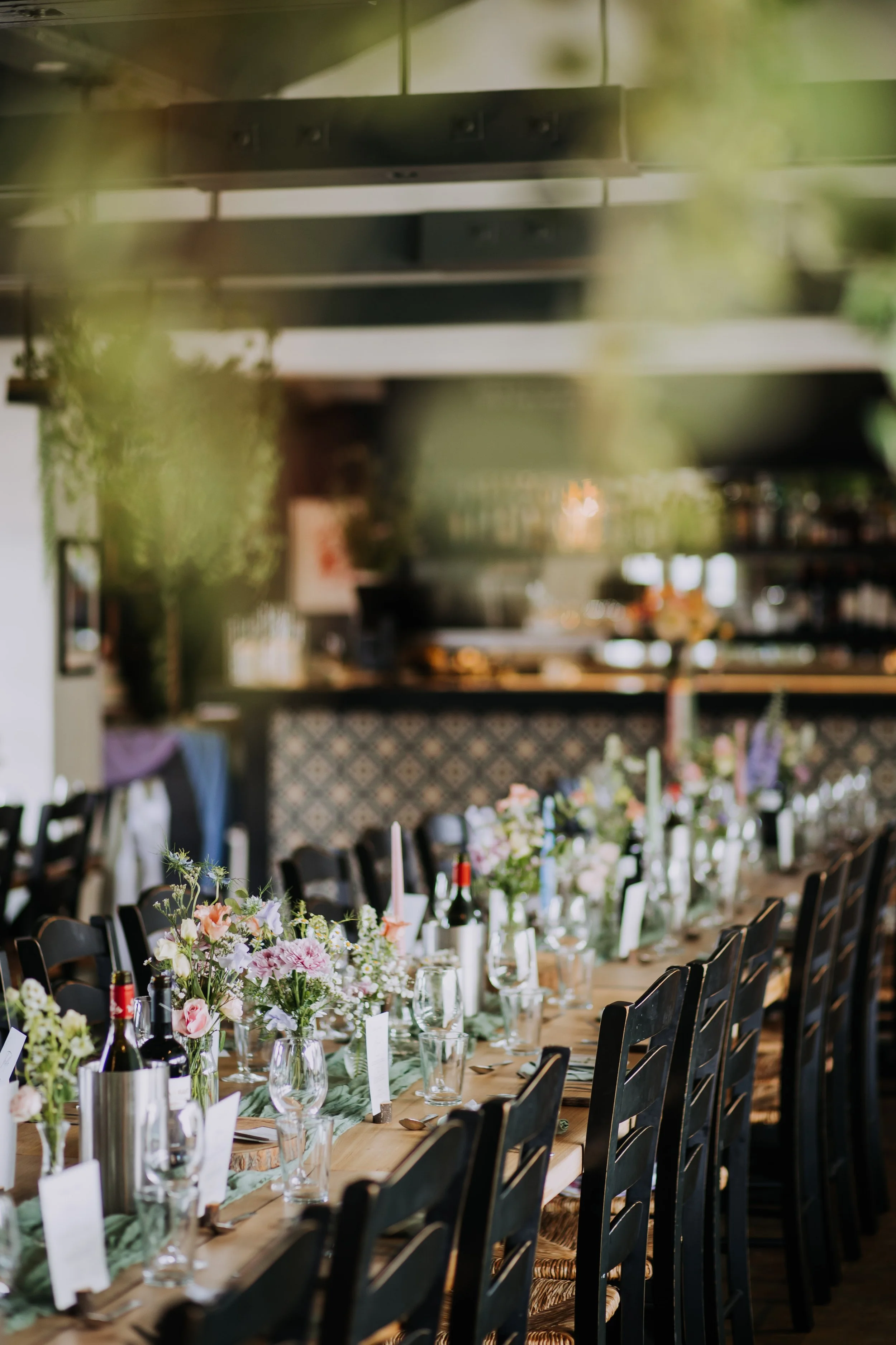 Long wooden dining table decorated with flowers, wine bottles, glasses, and candles in a restaurant or event space.