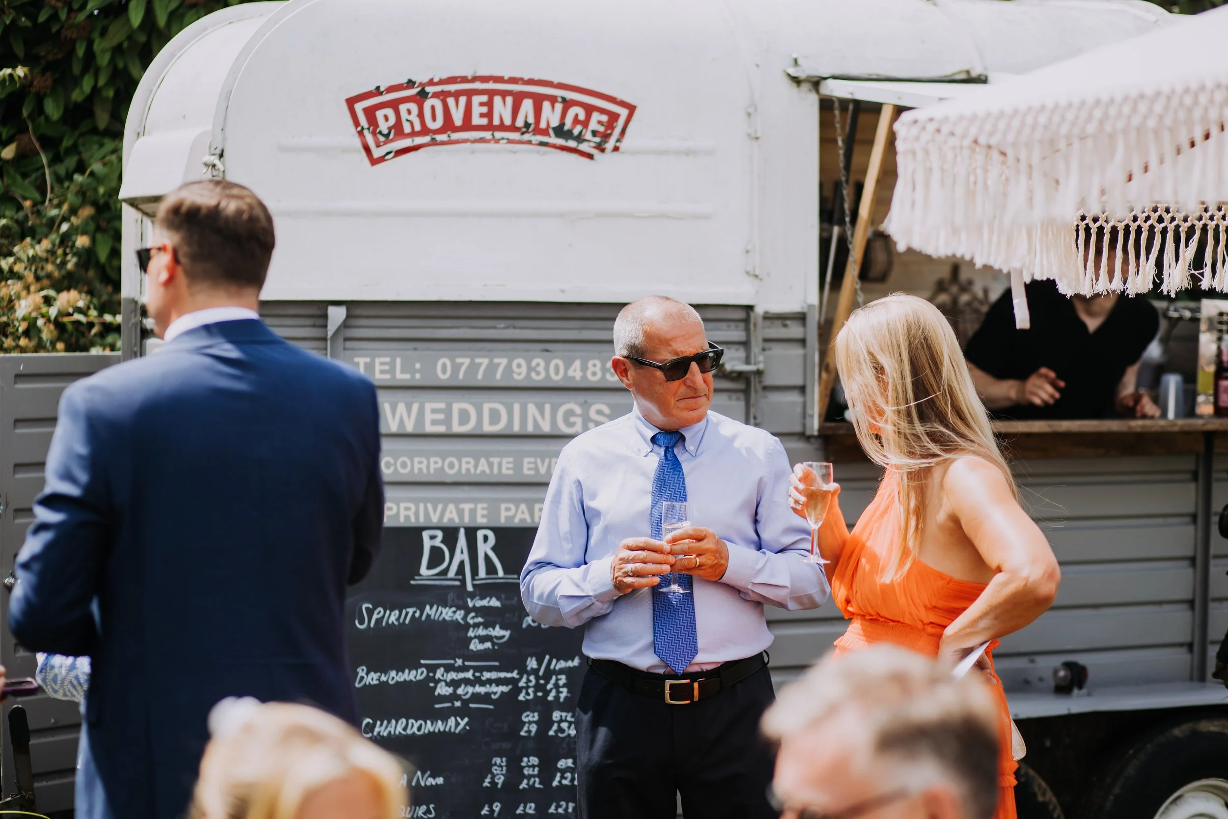 People socializing outdoors near a white mobile bar with a sign, in a festive event setting. One man with sunglasses and a woman in an orange dress are holding drinks and talking, while a man in a suit is seen from behind in the foreground.