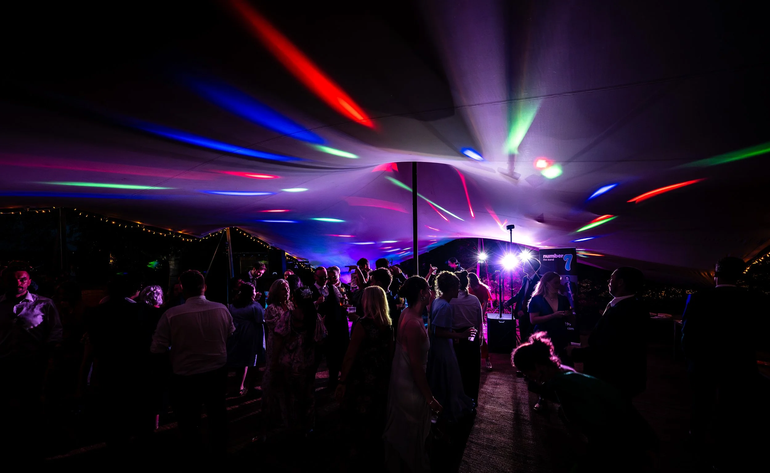 People dancing and socializing at a party under colorful stage lighting with a DJ booth and a tent ceiling.