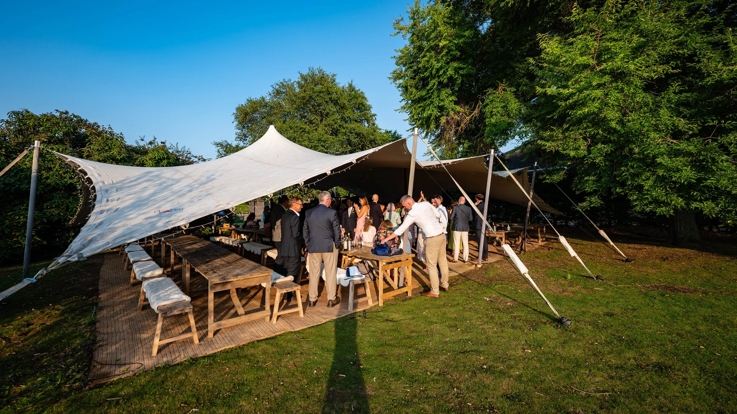 People gathered under a white canopy tent at an outdoor event in the late afternoon, with green trees and a clear blue sky in the background.