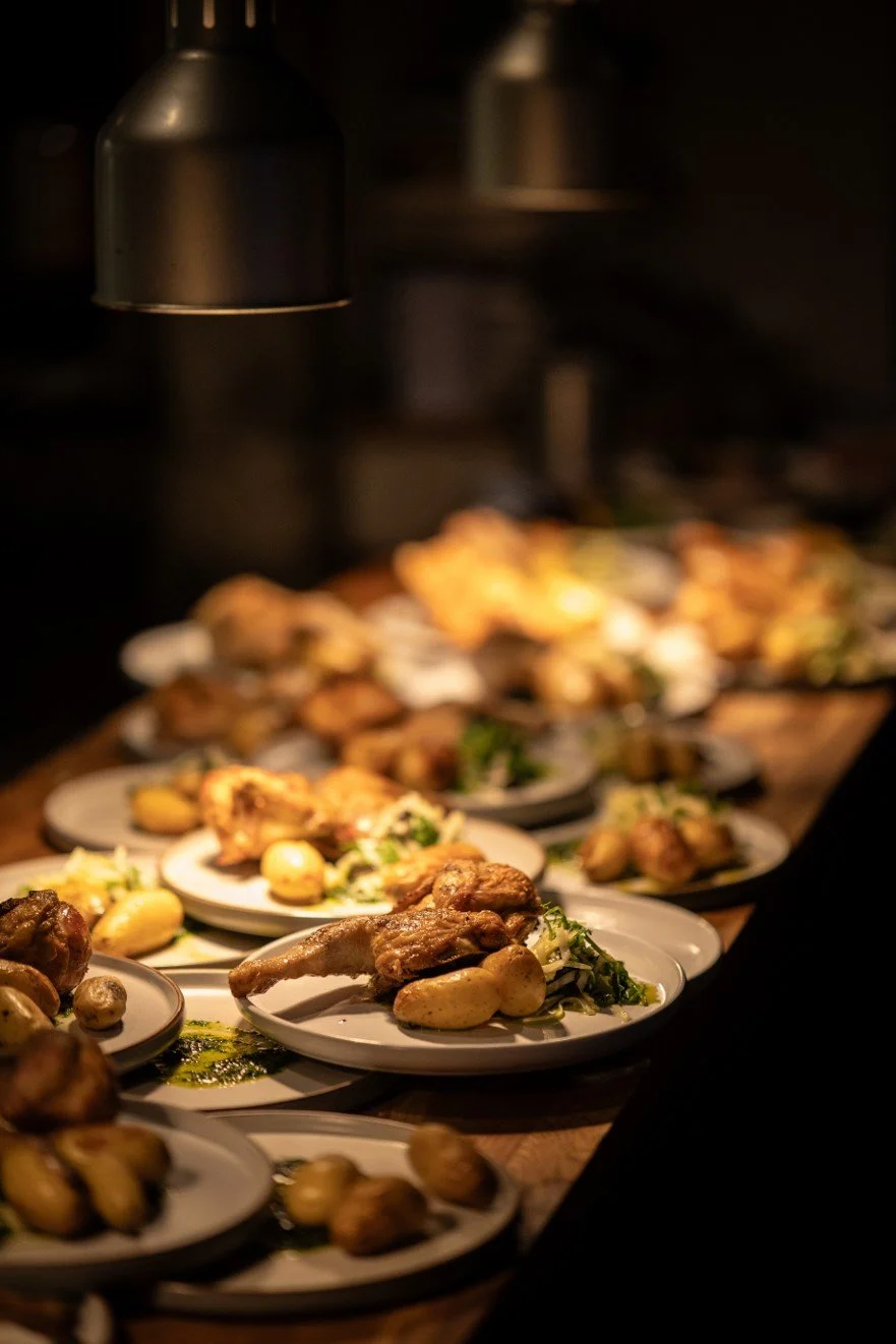 Multiple plates of roasted chicken, potatoes, and vegetables under warm lighting in a dark setting.