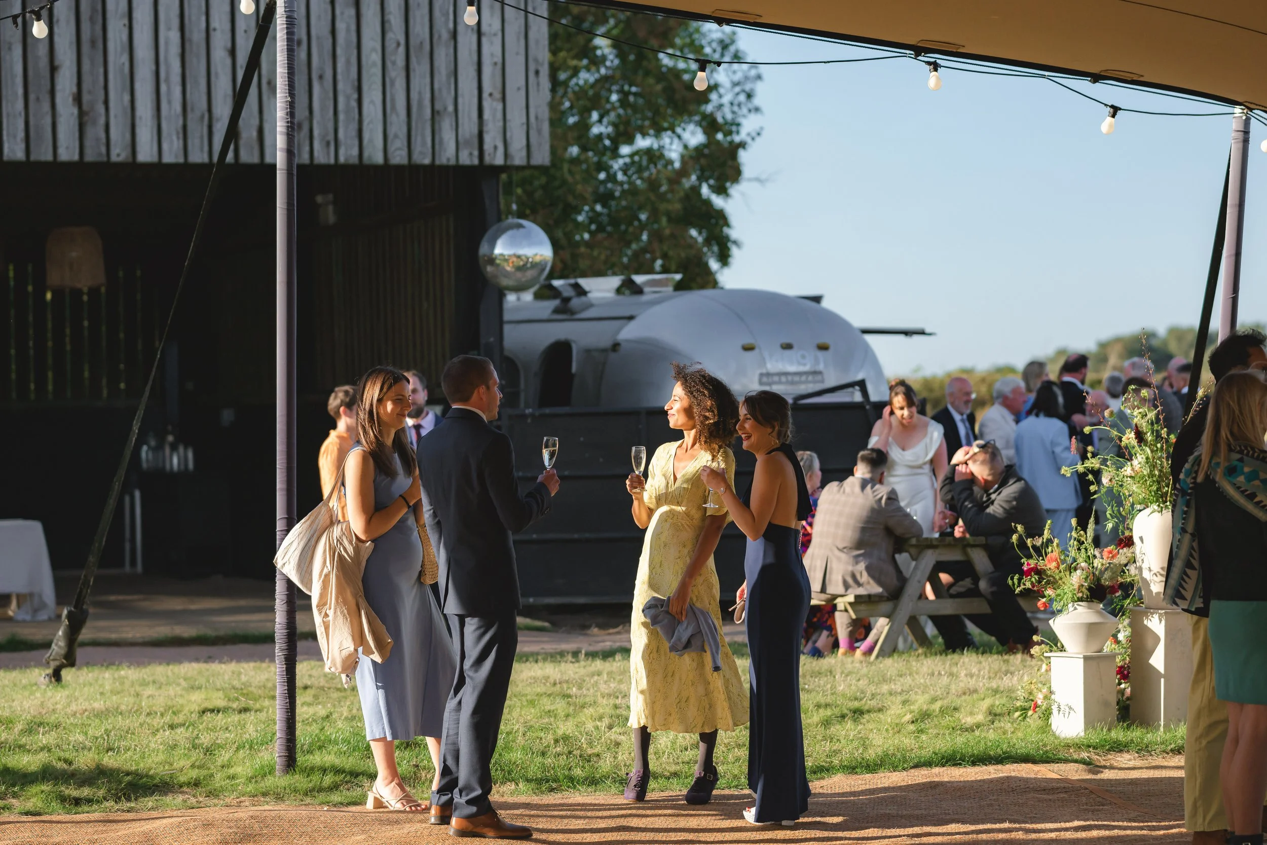 Guests at an outdoor wedding reception talk and drink champagne under a tent, with string lights and floral arrangements, and a vintage trailer in the background.