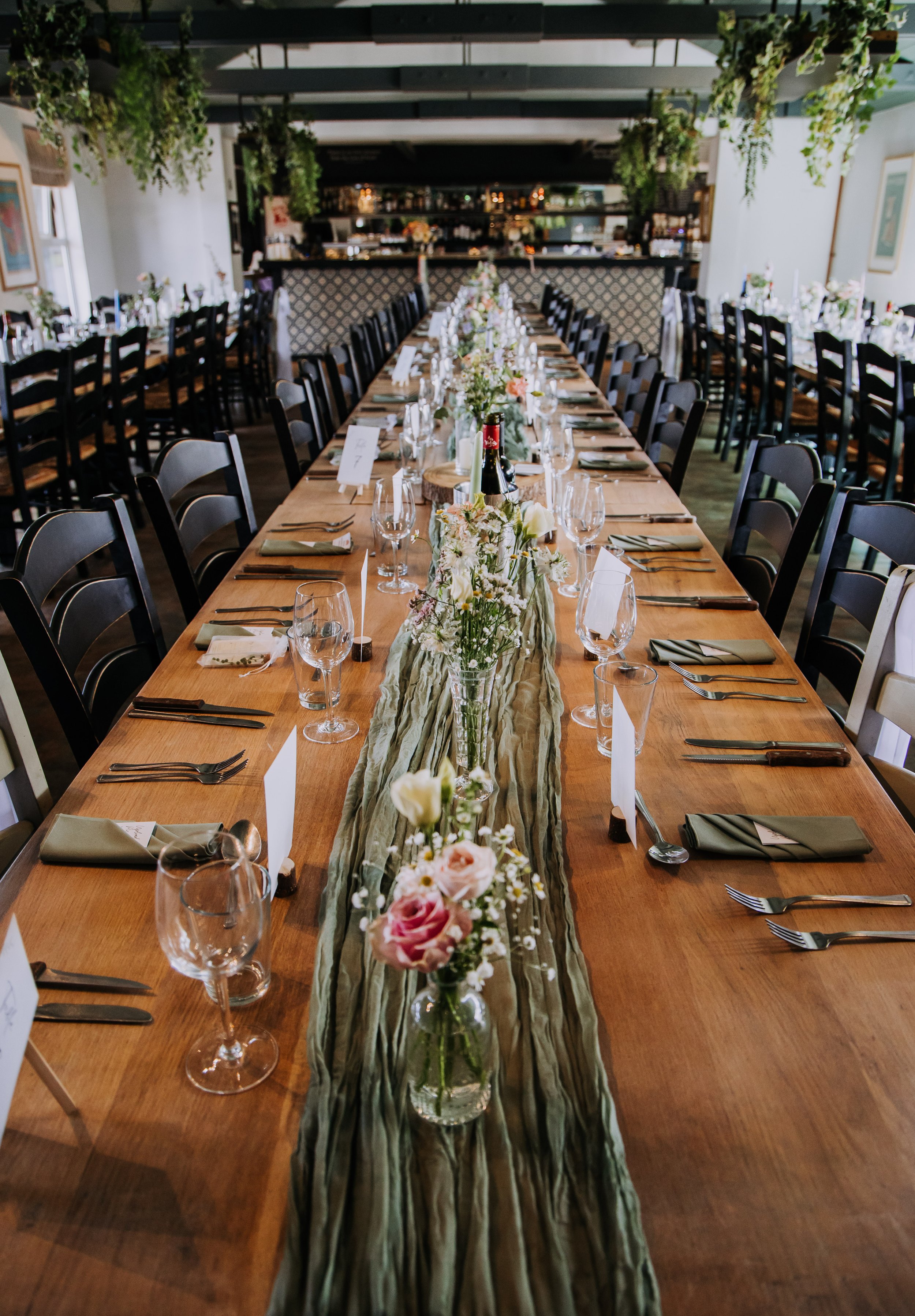 Long wooden dining table decorated with a floral centerpiece on a green fabric table runner, set with wine glasses, water glasses, forks, knives, and cloth napkins, in a well-lit room with hanging greenery and framed artwork.