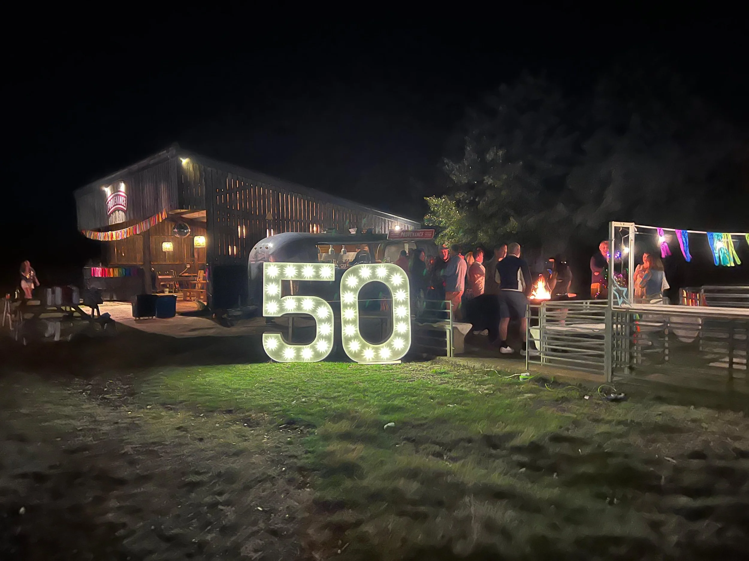 Nighttime outdoor celebration with illuminated number 50, people gathered around a fire pit, a rustic wooden building with decorations, and colorful banners.