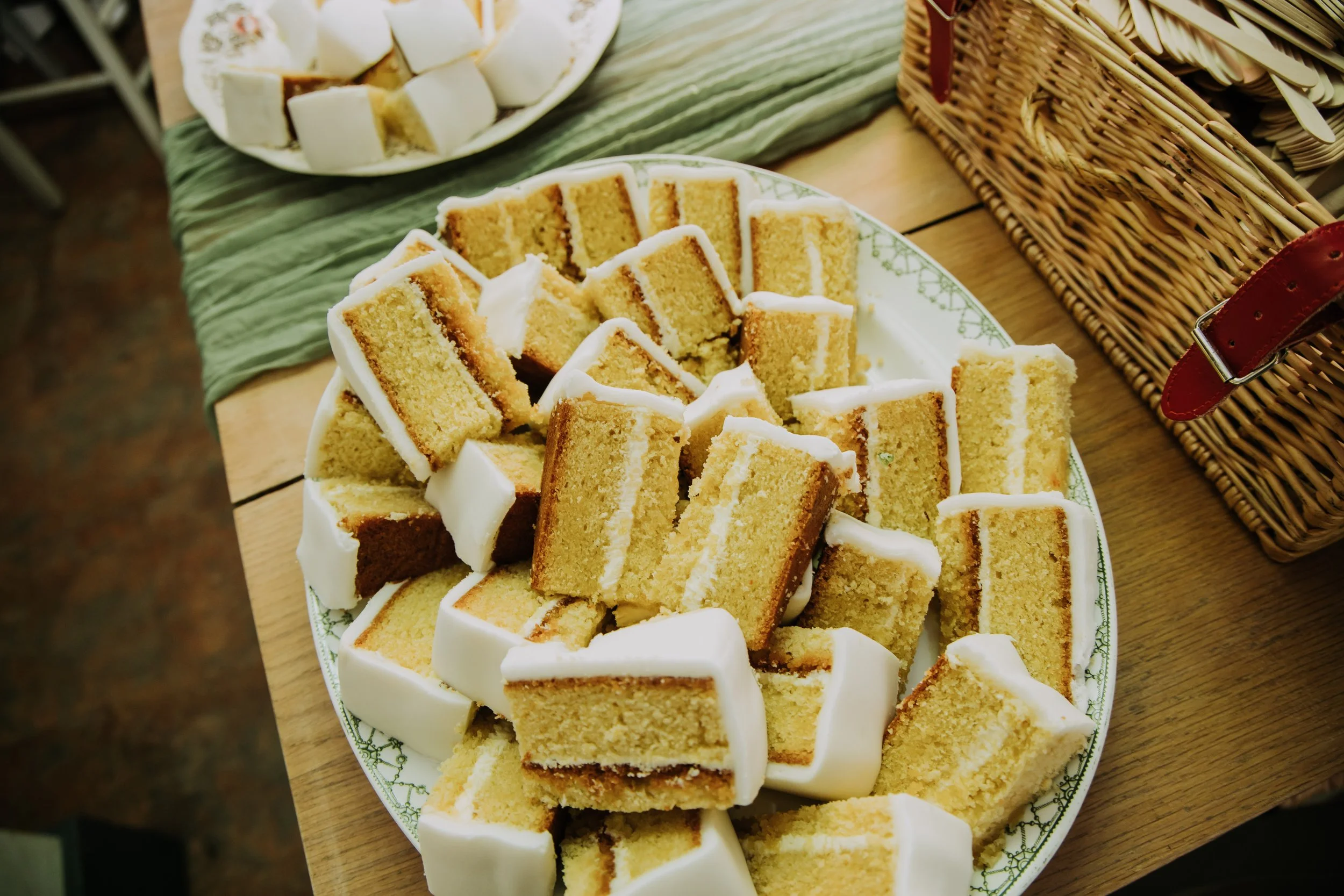 A plate of sliced vanilla cake with white frosting on a wooden table, with a green cloth underneath and a wicker basket nearby.
