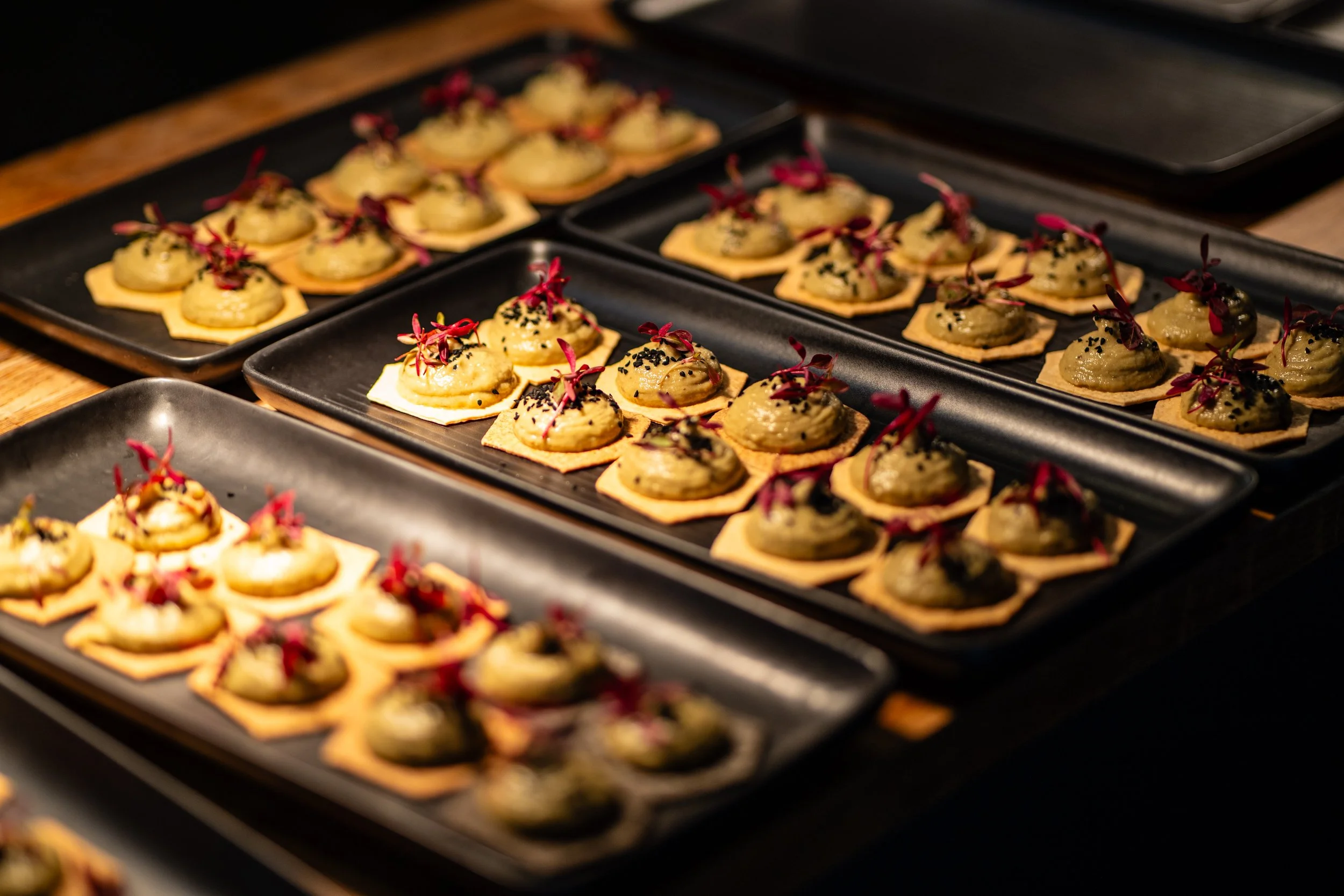 Multiple black serving trays filled with small appetizers consisting of crispy crackers topped with a creamy spread, garnished with red microgreens and black sesame seeds.