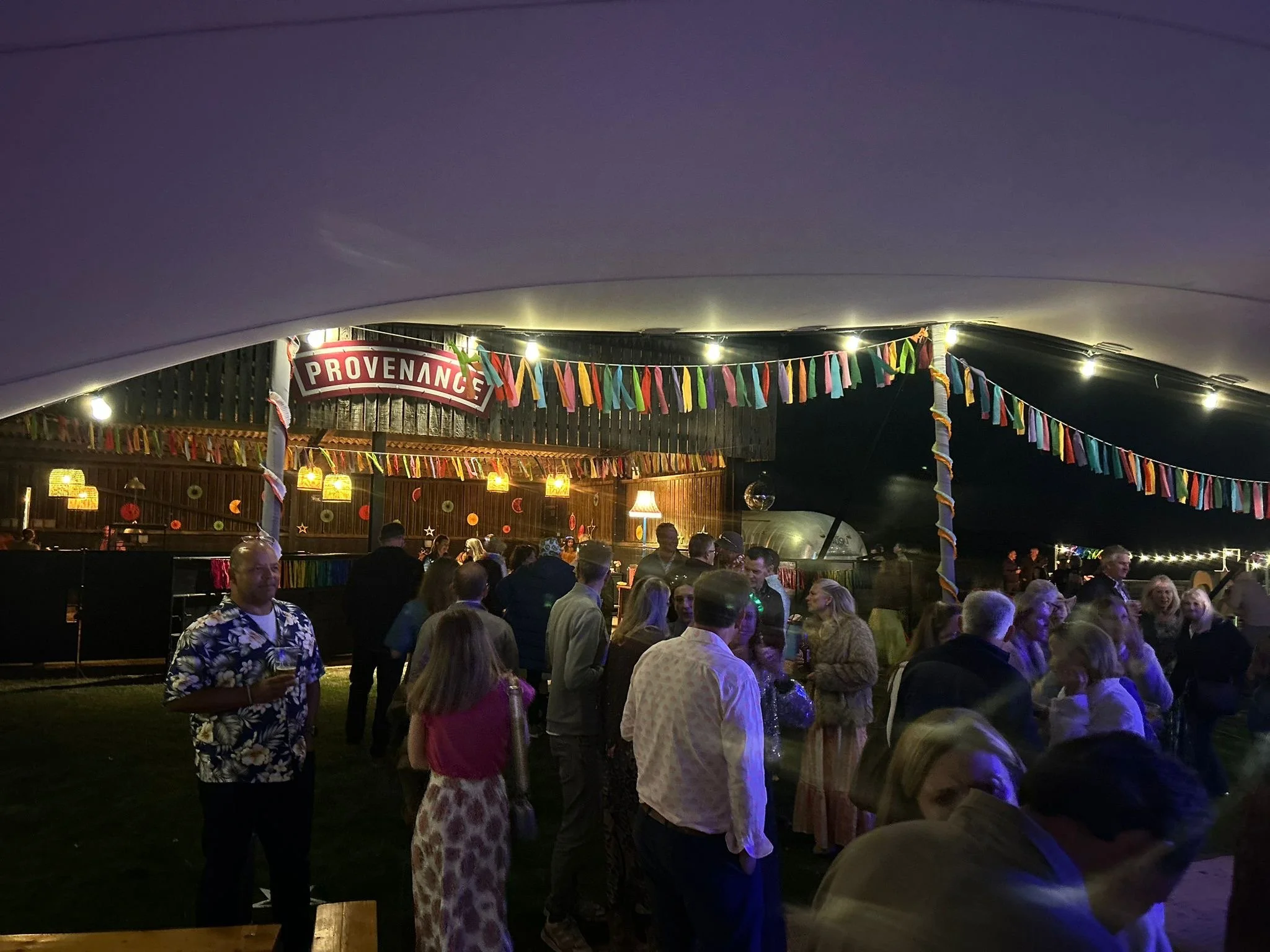 Nighttime outdoor party with colorful decorations, string lights, and a crowd of people socializing near a bar area under a large tent.