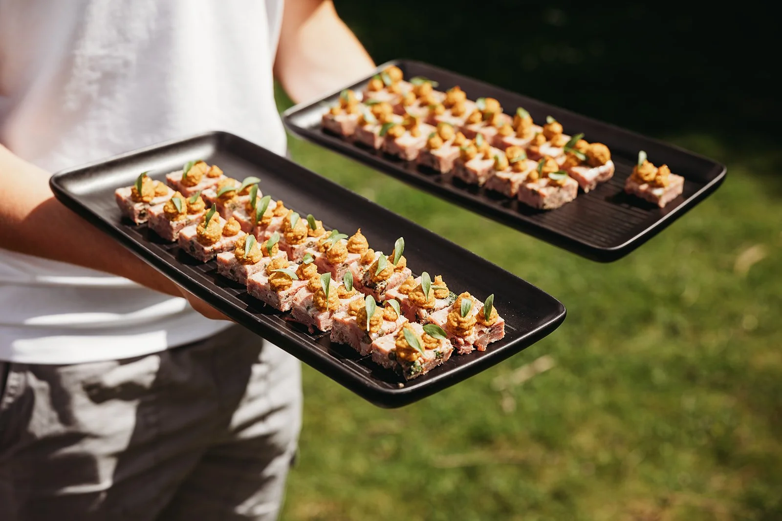 Person holding two black trays with small rectangular snacks topped with nuts and green herbs outdoors.