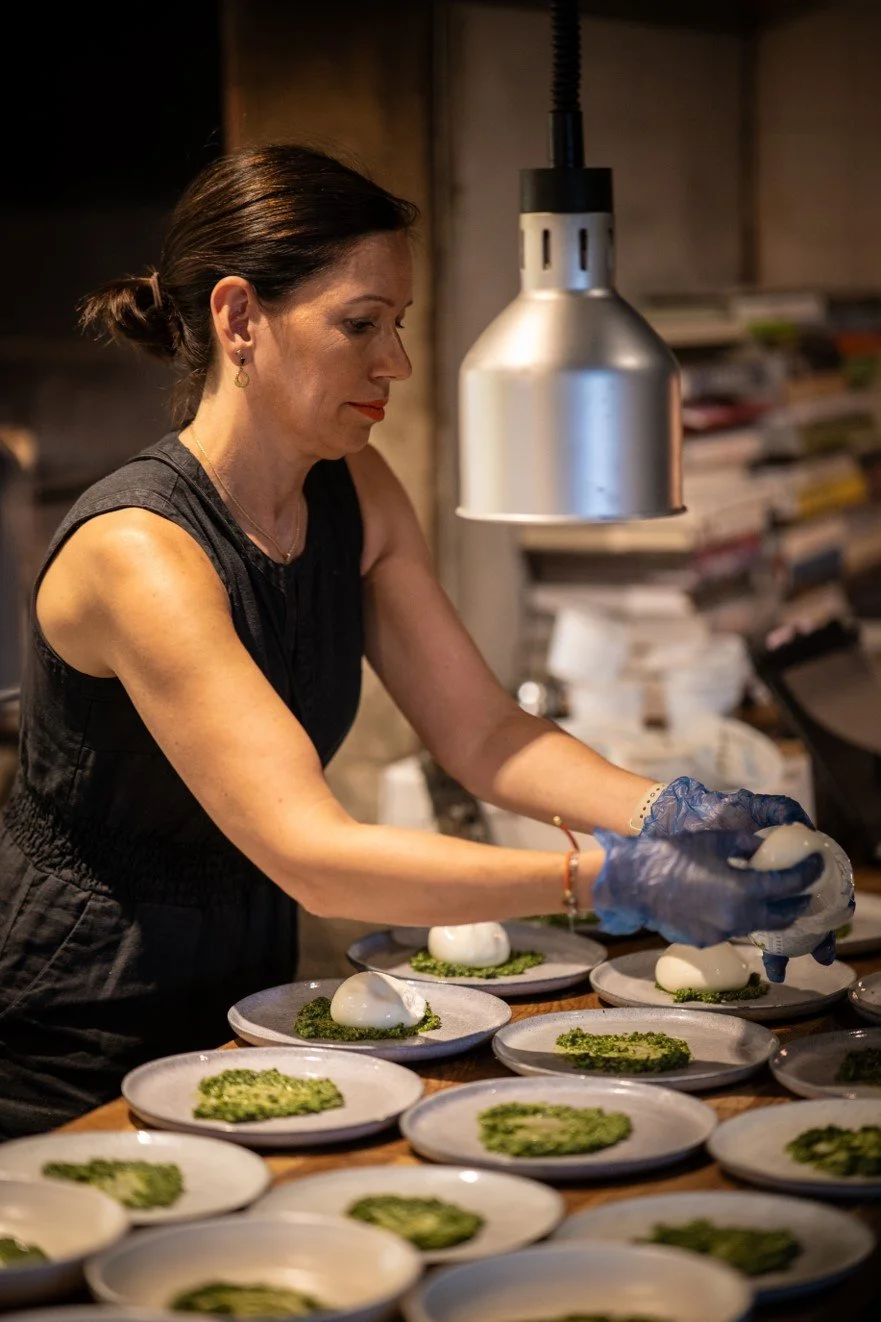 A woman wearing a black sleeveless top and blue gloves preparing multiple dishes with green garnish in a kitchen.