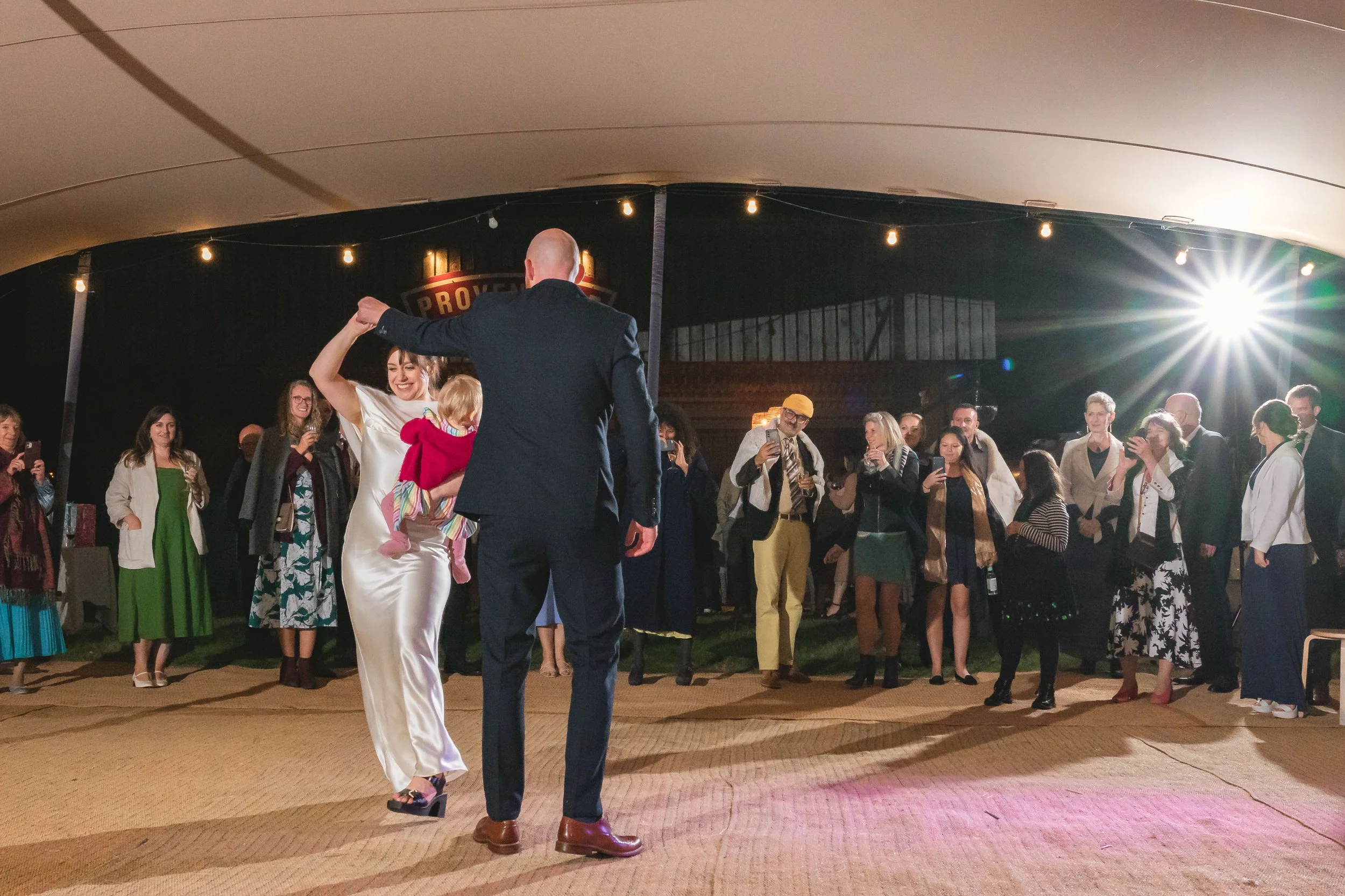 A woman in a white satin dress dancing with a man in a dark suit, holding a small child, at a social event with many guests watching and taking photos, under a decorative tent with string lights.