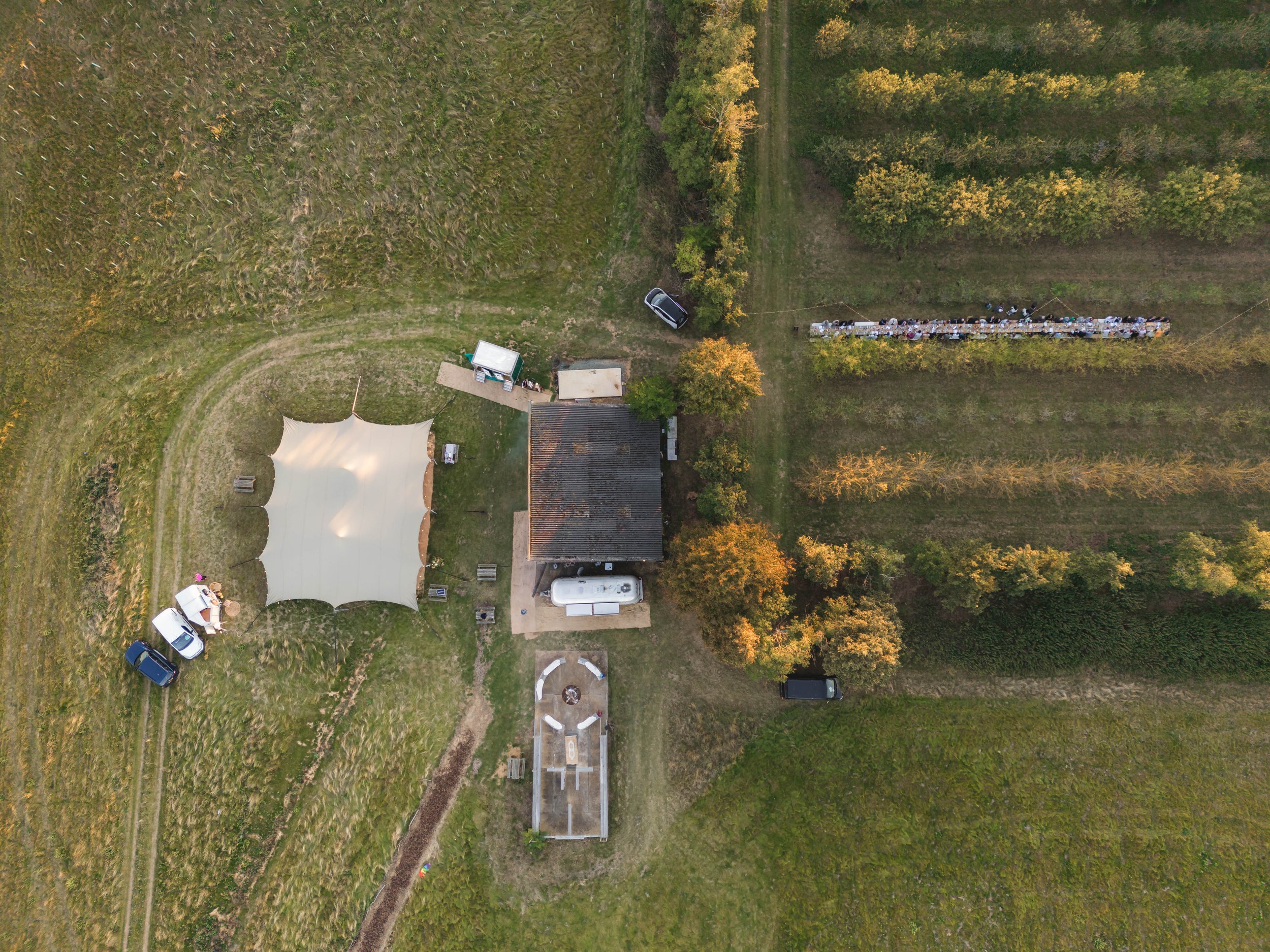 An aerial view of a countryside scene featuring a small building with a dark roof, a large white canopy tent, several parked cars, a picnic area with tables, and surrounding fields and trees, some showing fall foliage.